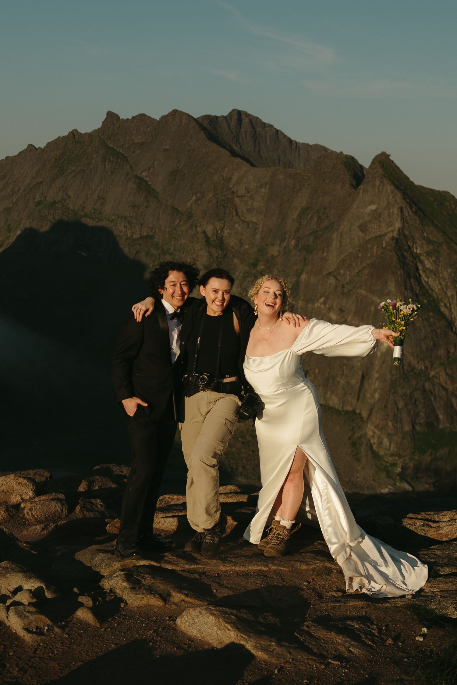 Two brides and photographer pose joyfully on rocky mountain summit during golden hour elopement ceremony.