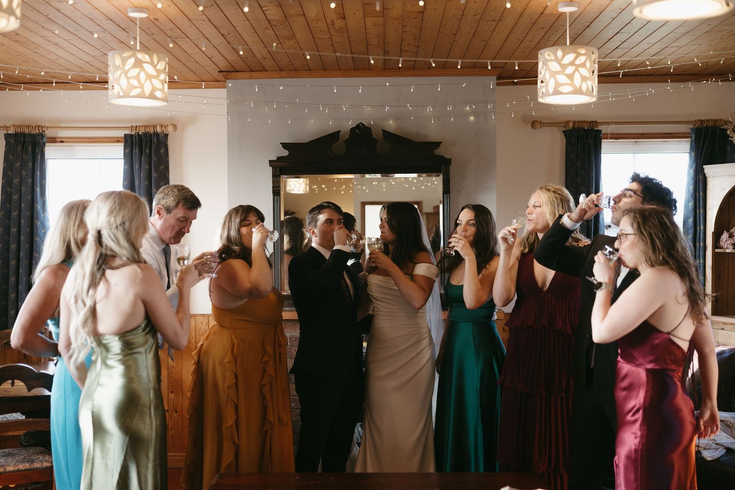 Wedding guests dance and celebrate together on a wooden-floored reception venue with pendant lighting and rustic decor.