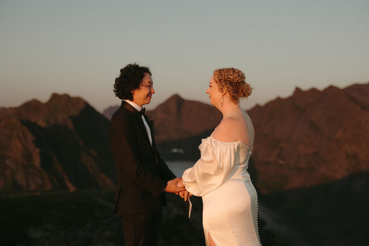 Newlywed couple embraces in romantic silhouette against dramatic mountain landscape at sunset.
