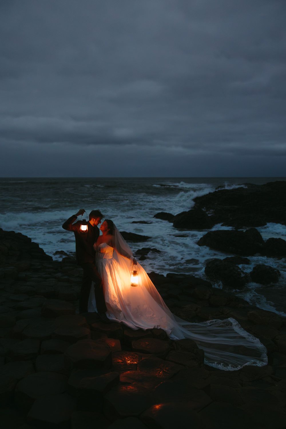 Lantern-lit wedding photos on rocky shore at night with crashing waves.