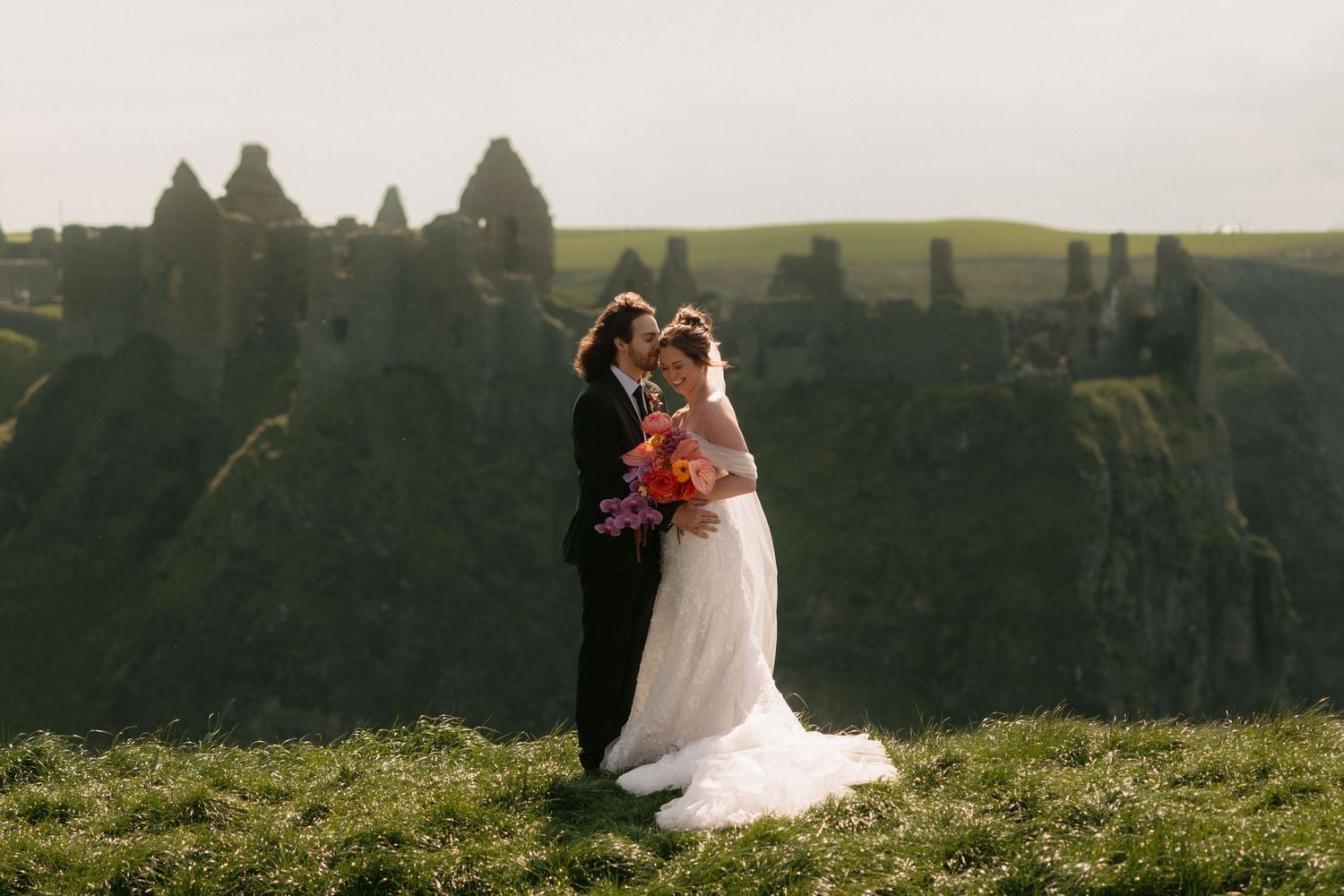 Couple sits together on grassy hilltop overlooking scenic mountain landscape during sunset.