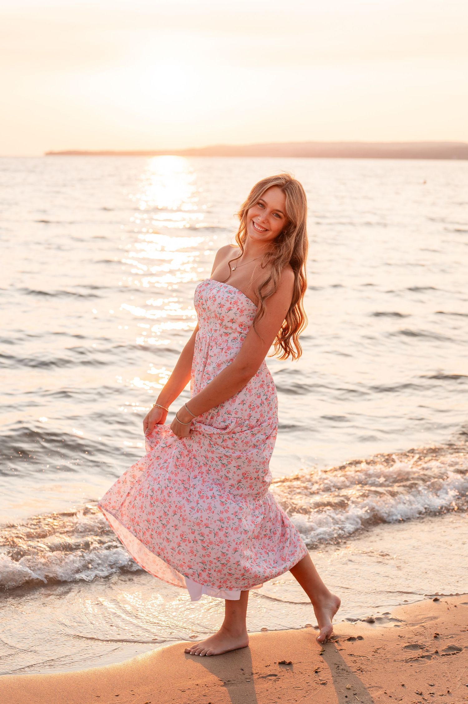 A senior girl in a floral dress twirls joyfully on the beach at sunset, creating a playful and carefree moment on Lake Michigan in Petoskey.