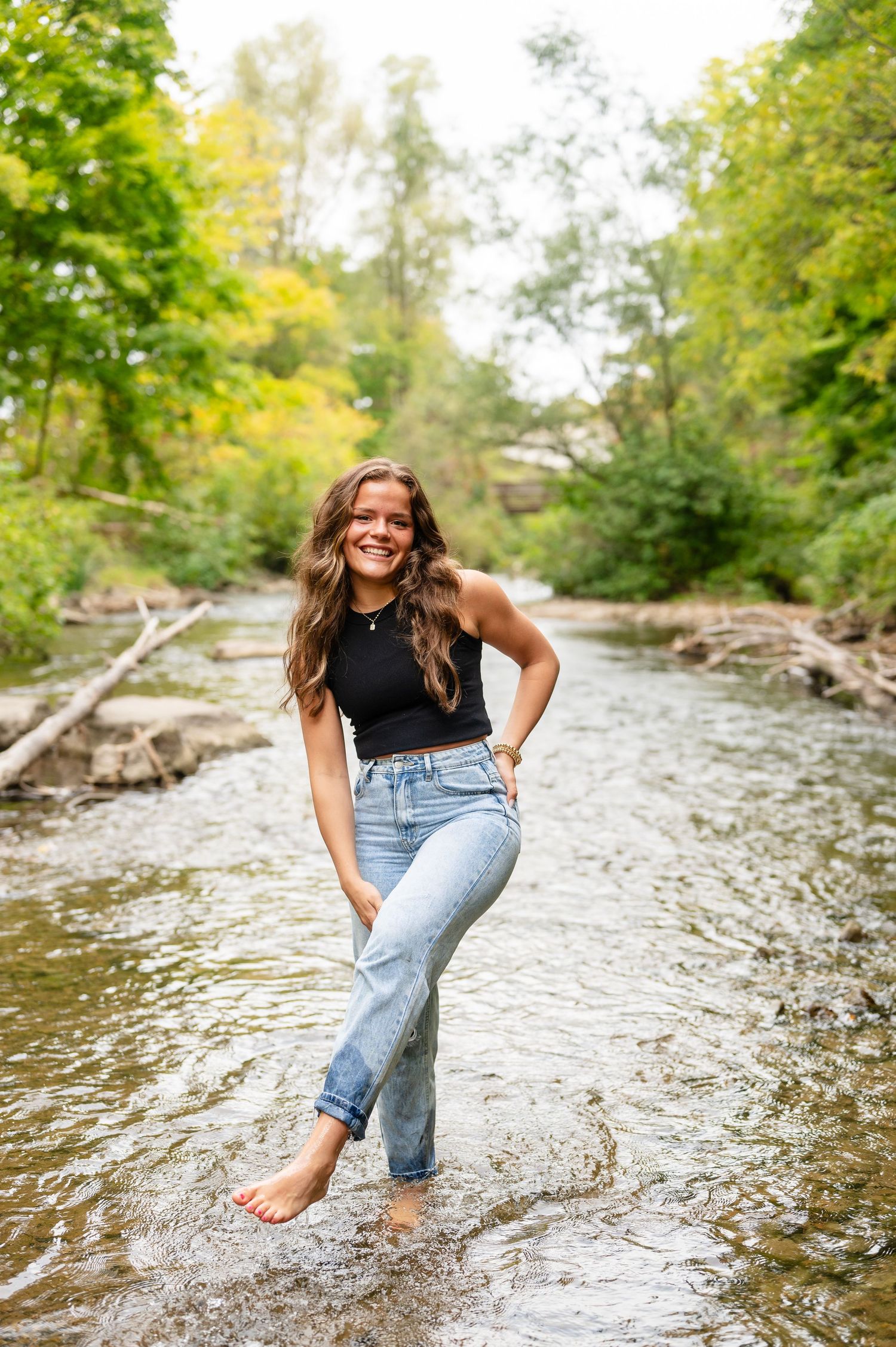 A casual outdoor portrait of a senior girl standing barefoot in a river wearing jeans and a black top.