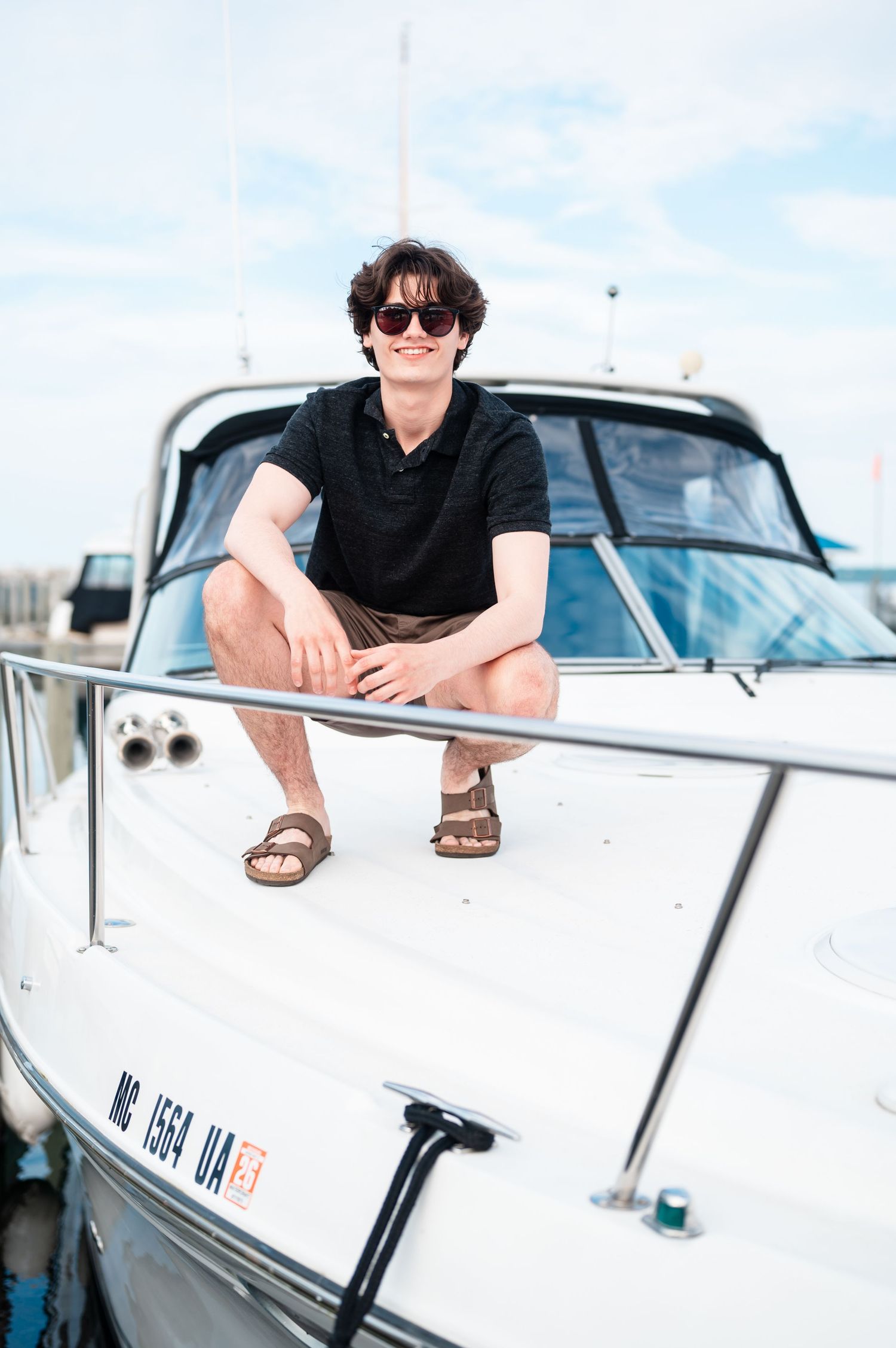 Senior boy relaxing on a white boat with sunglasses on against a bright sky background.