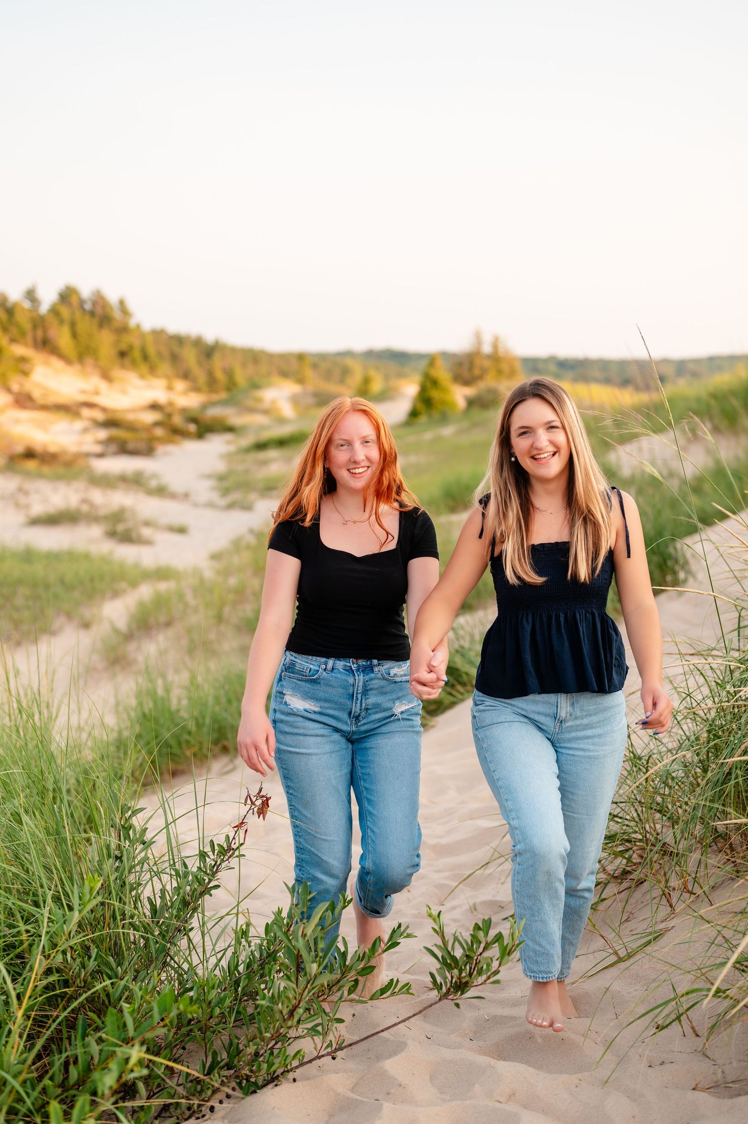 Two sisters walking together on sandy beach dunes with grass at sunset.