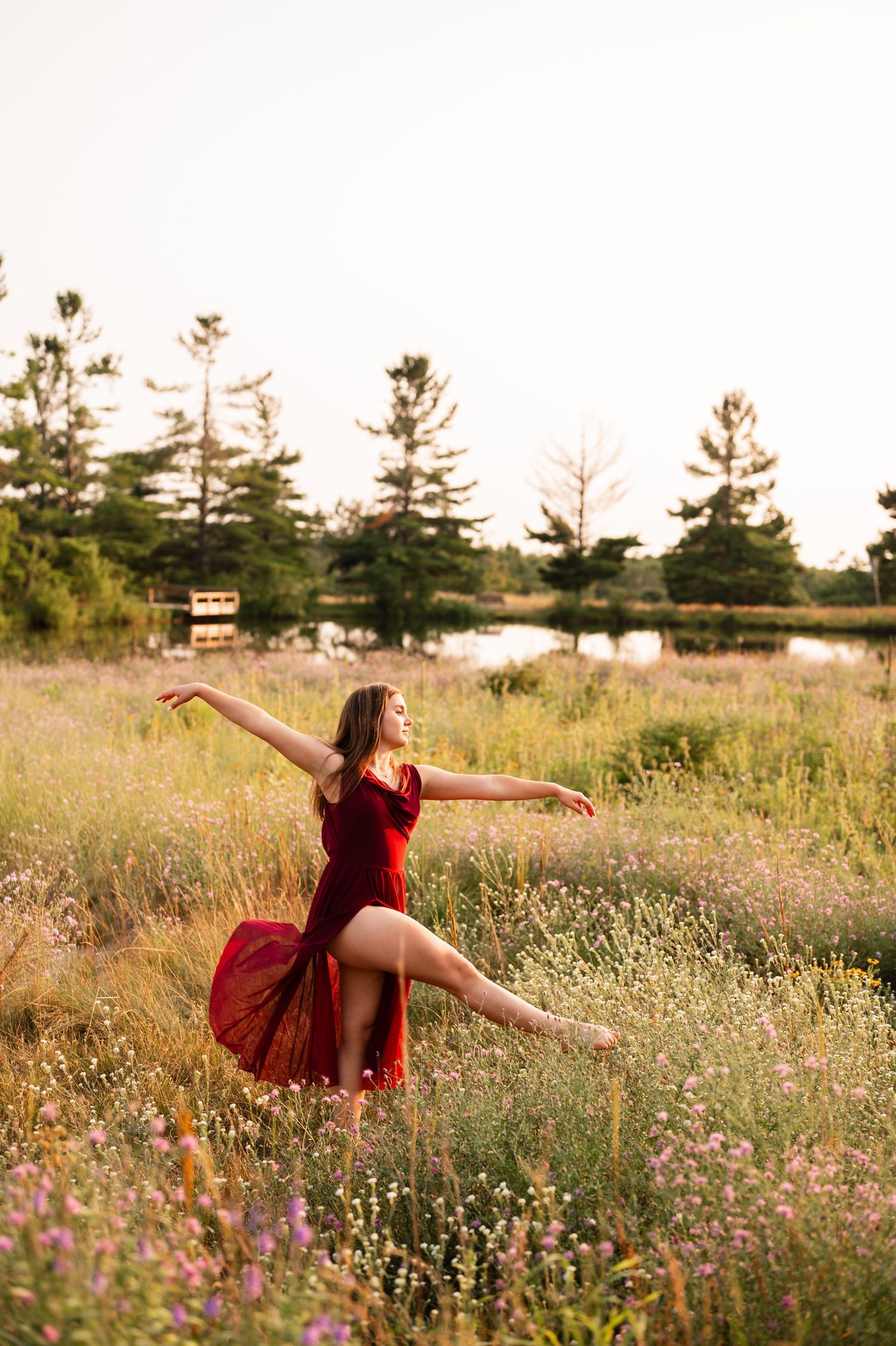 A dancer in a red dress performs gracefully in a wildflower field at golden hour with trees and a pond behind her.