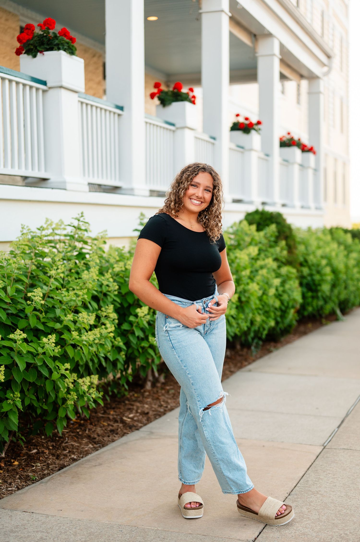 A casual portrait by a white columned porch in Petoskey Michigan with red flowers, wearing jeans and a black top.