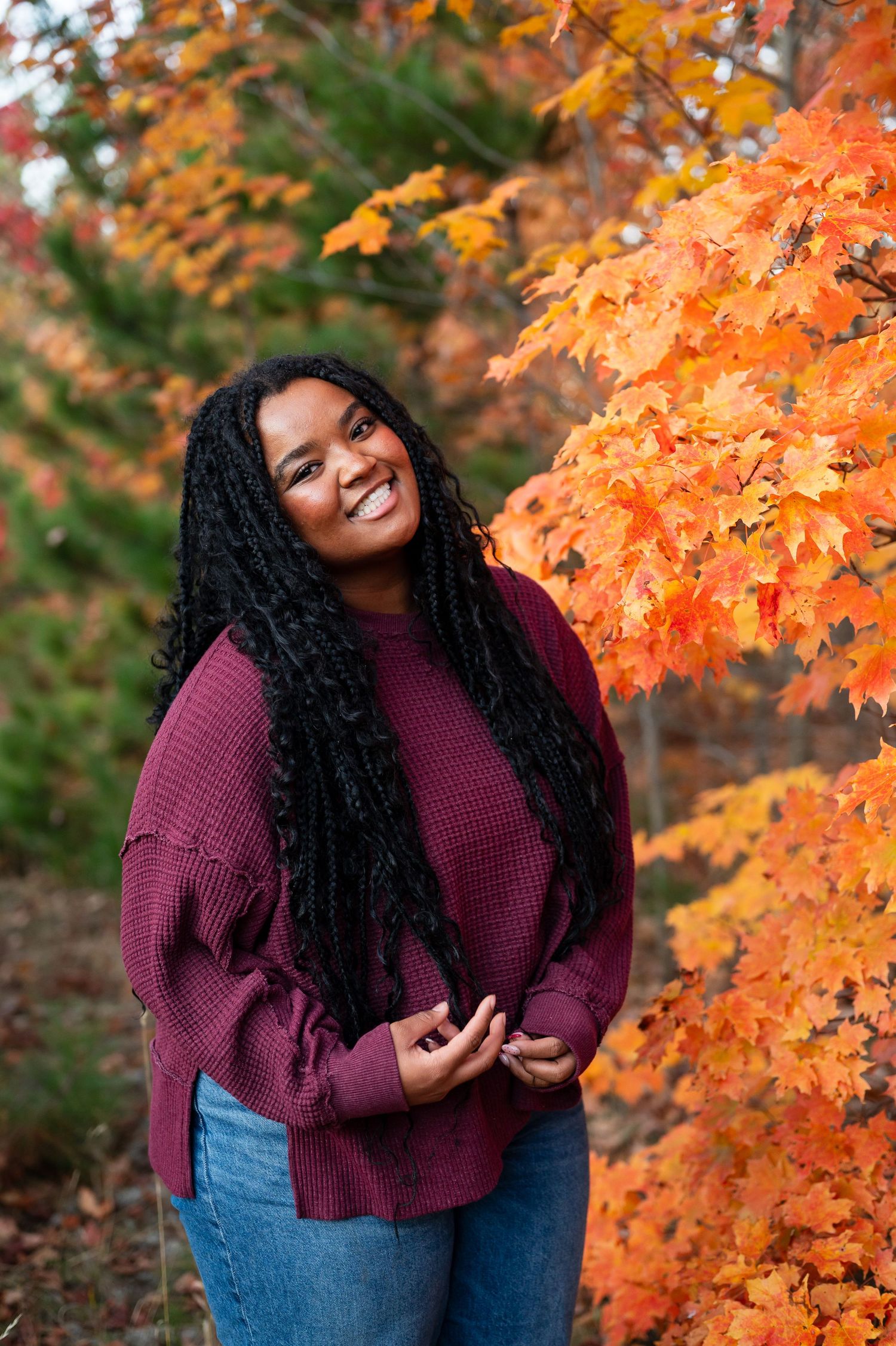 A senior girl in a burgundy sweater smiles brightly against vibrant orange autumn foliage in Gaylord, MI