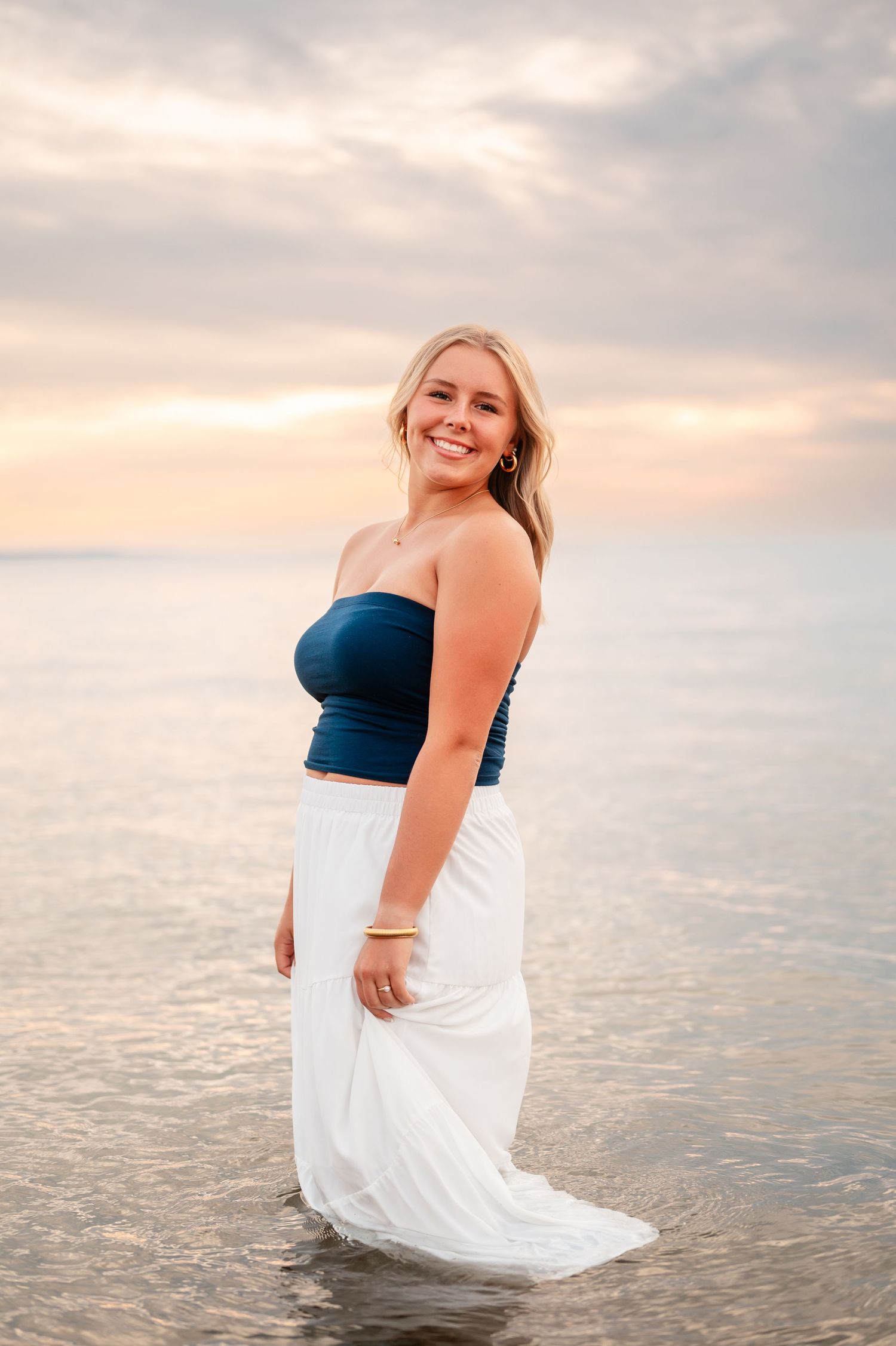 A senior girl in a navy top and white skirt poses gracefully by the water during sunset.