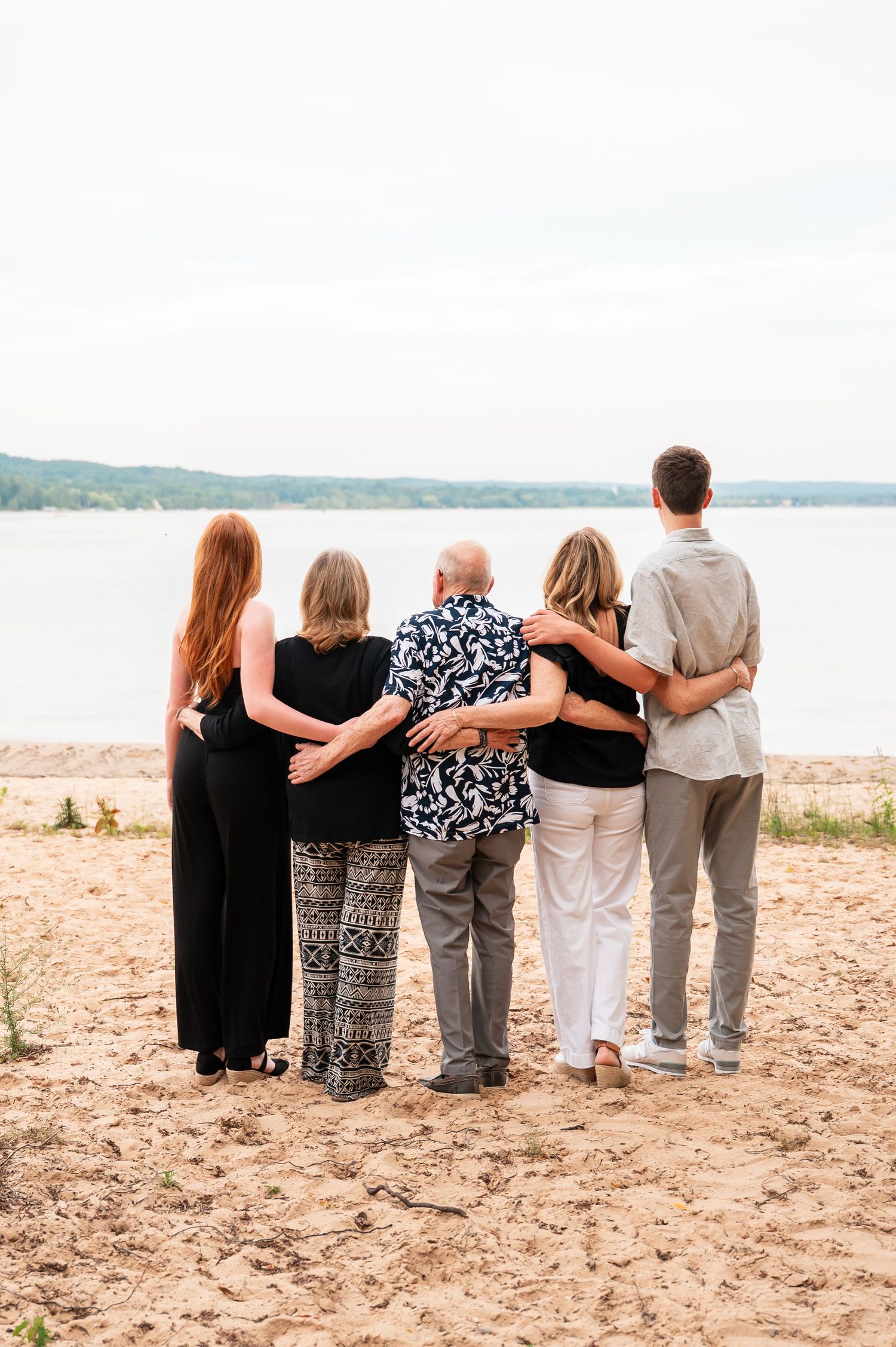 Group stands together with arms around each other on a sandy beach overlooking water.