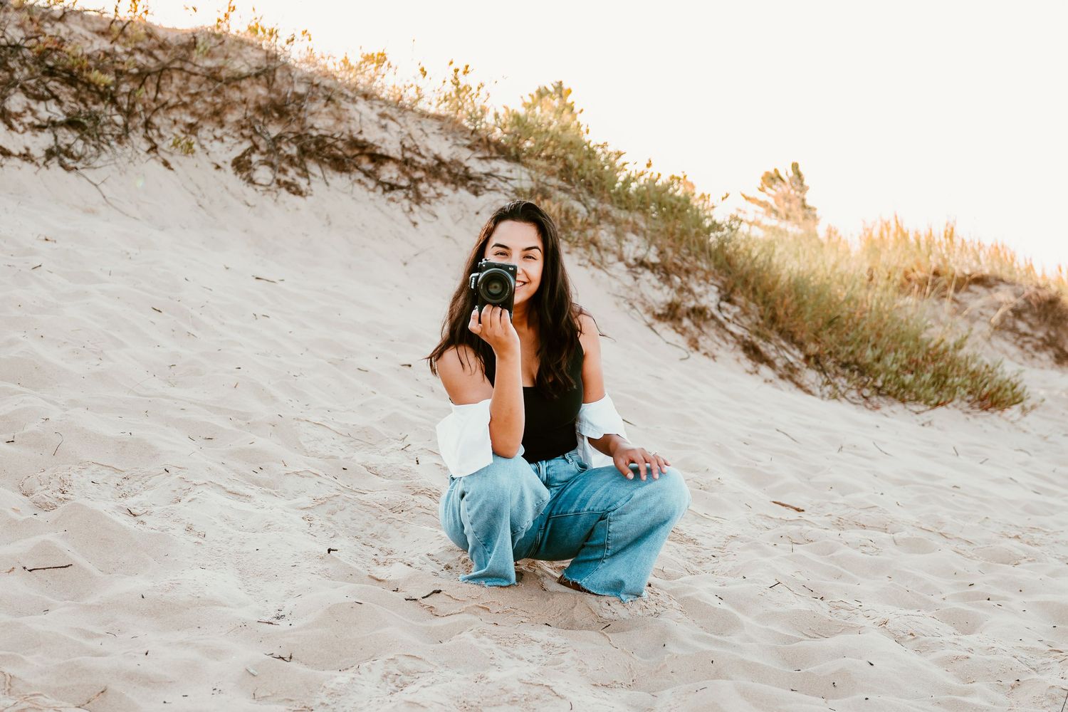 A photographer with camera squatting on sandy beach with jeans.
