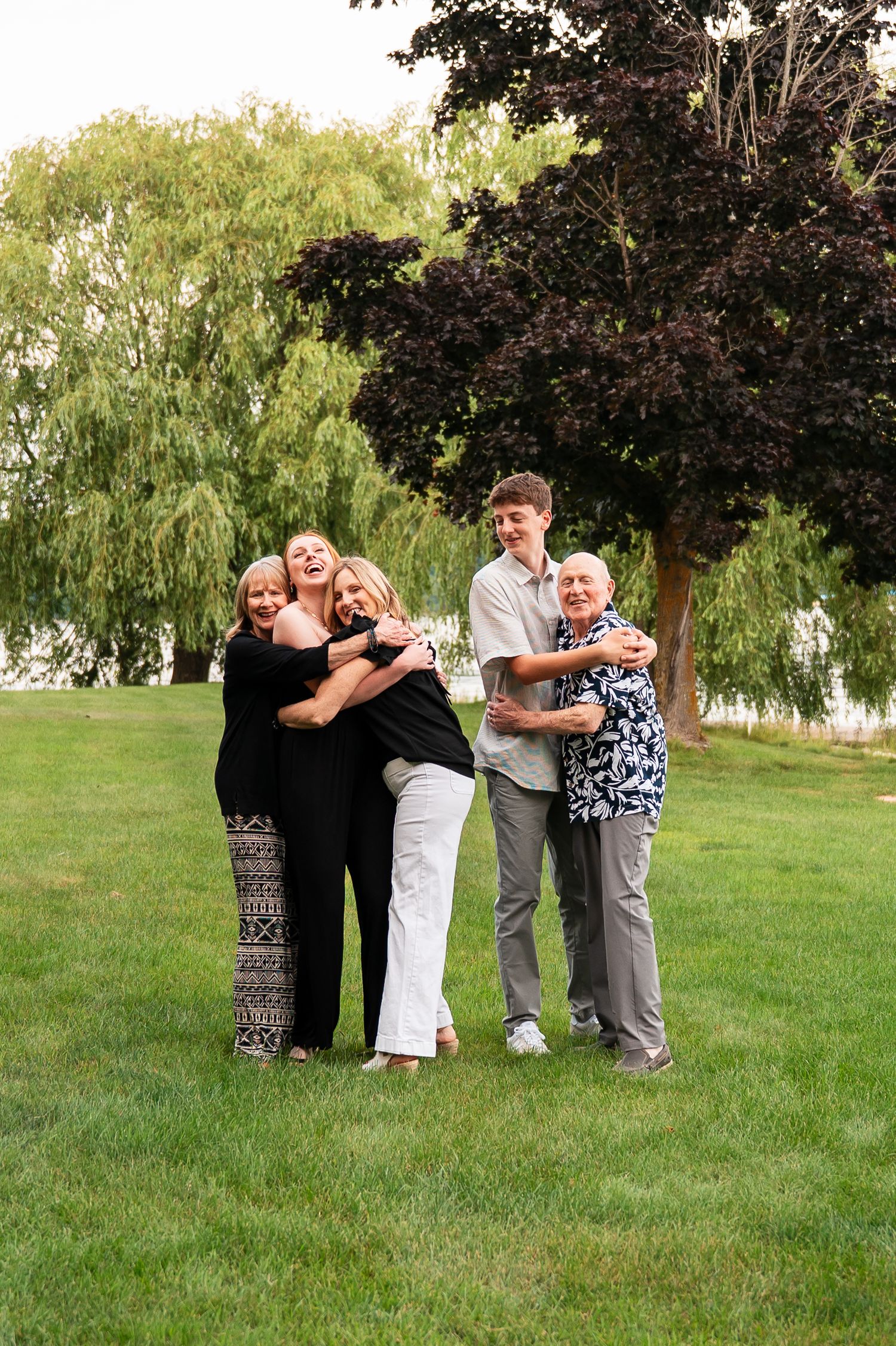 Family hugs on green lawn with willow trees in background.