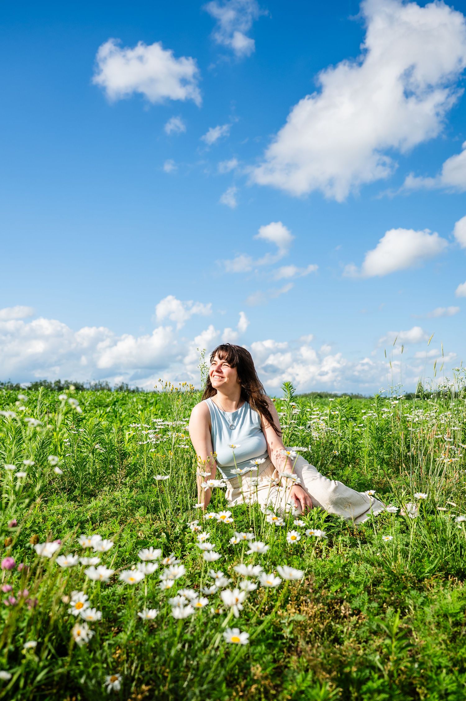 A girl in white pants sits among blooming daisies in a sunny meadow under blue skies. and clouds in Northern Michiga.