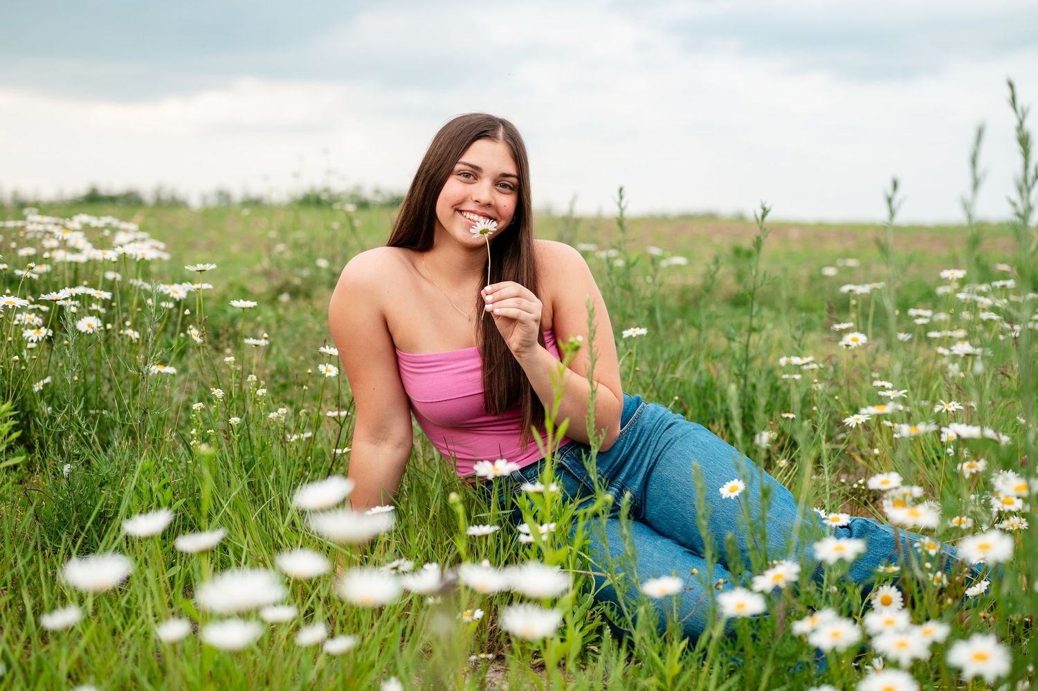 Senior girl relaxing among blooming daisies in summer meadow wearing pink top and jeans in Gaylord Northern Michigan.