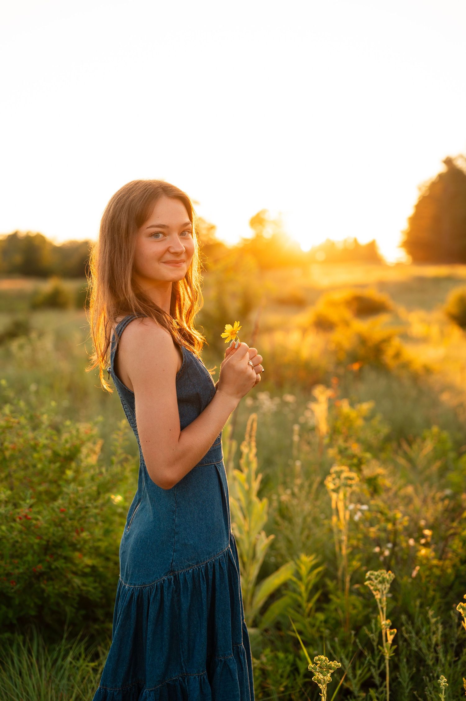 Young woman in teal dress holding wildflowers in a sunlit meadow at golden hour sunset.