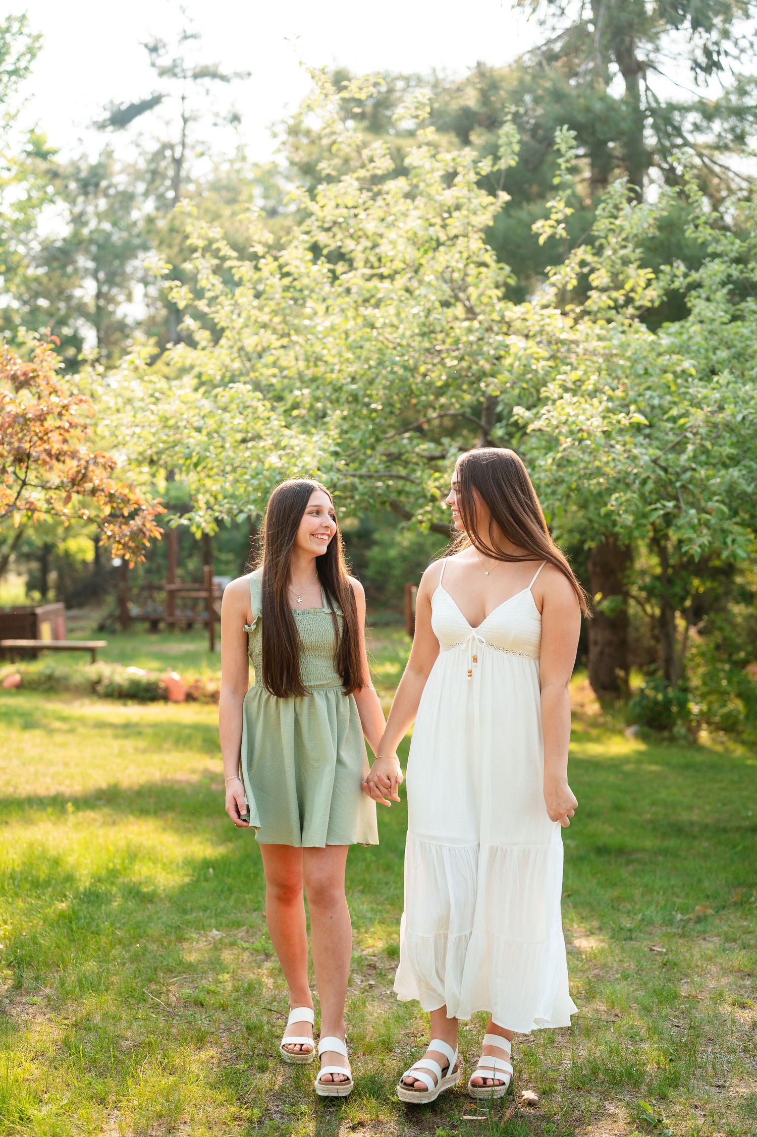 Two sisters in summer dresses hold hands while walking through a sunlit garden path.