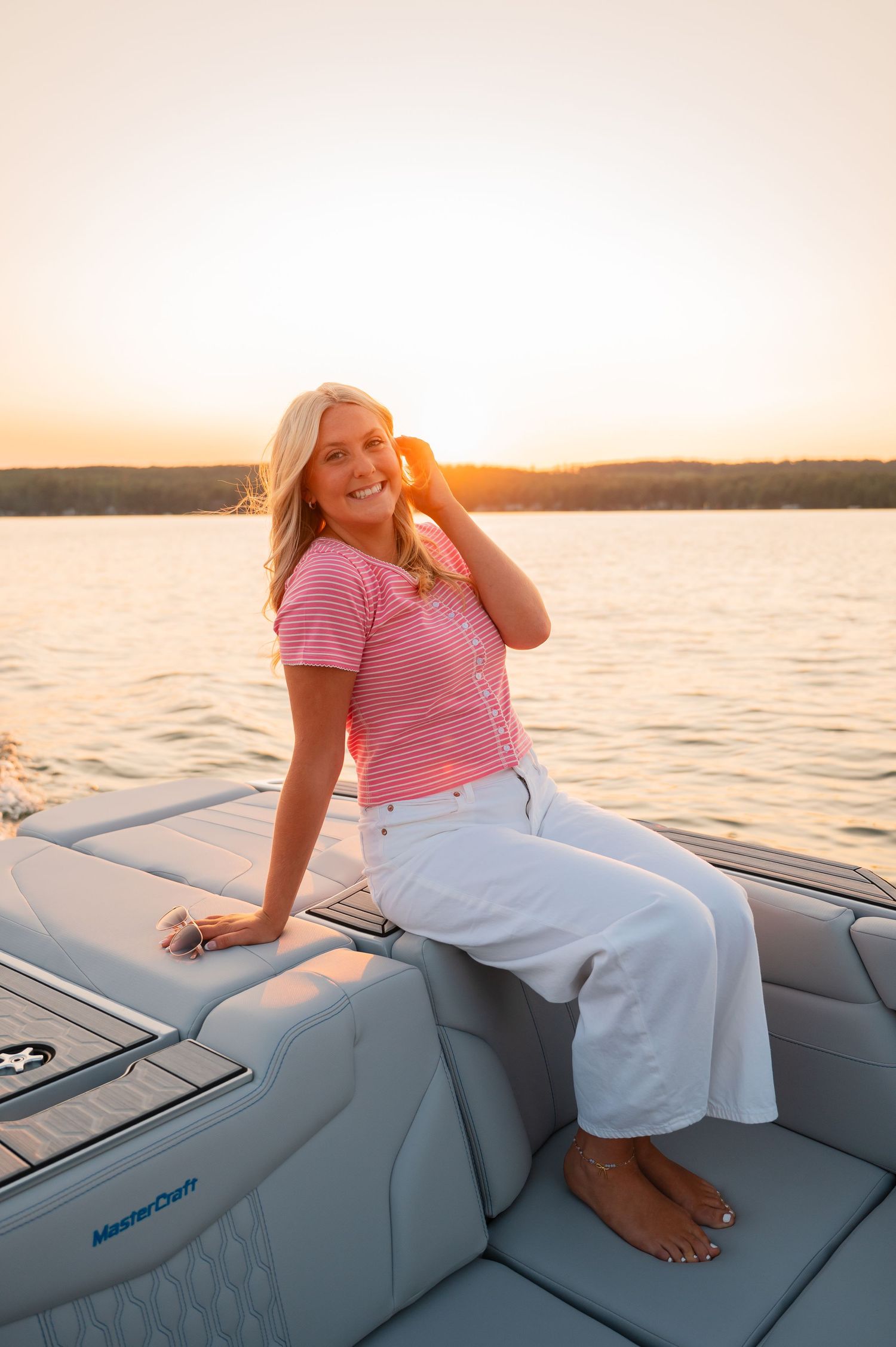 Blonde woman in pink striped top and white pants sitting on a MasterCraft boat at golden hour sunset.