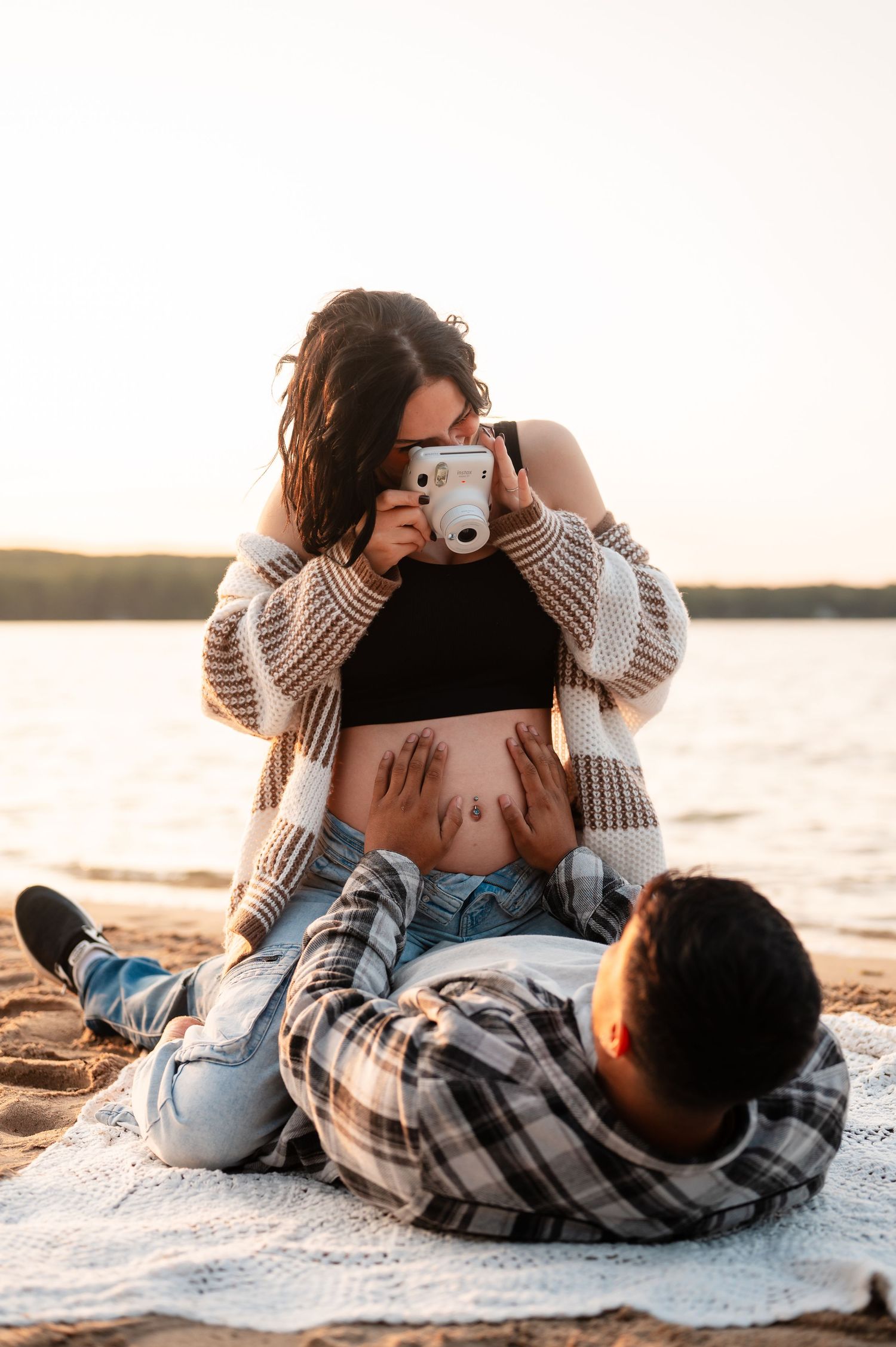 A maternity session with a couple relaxing on a blanket at the beach during golden hour sunset.