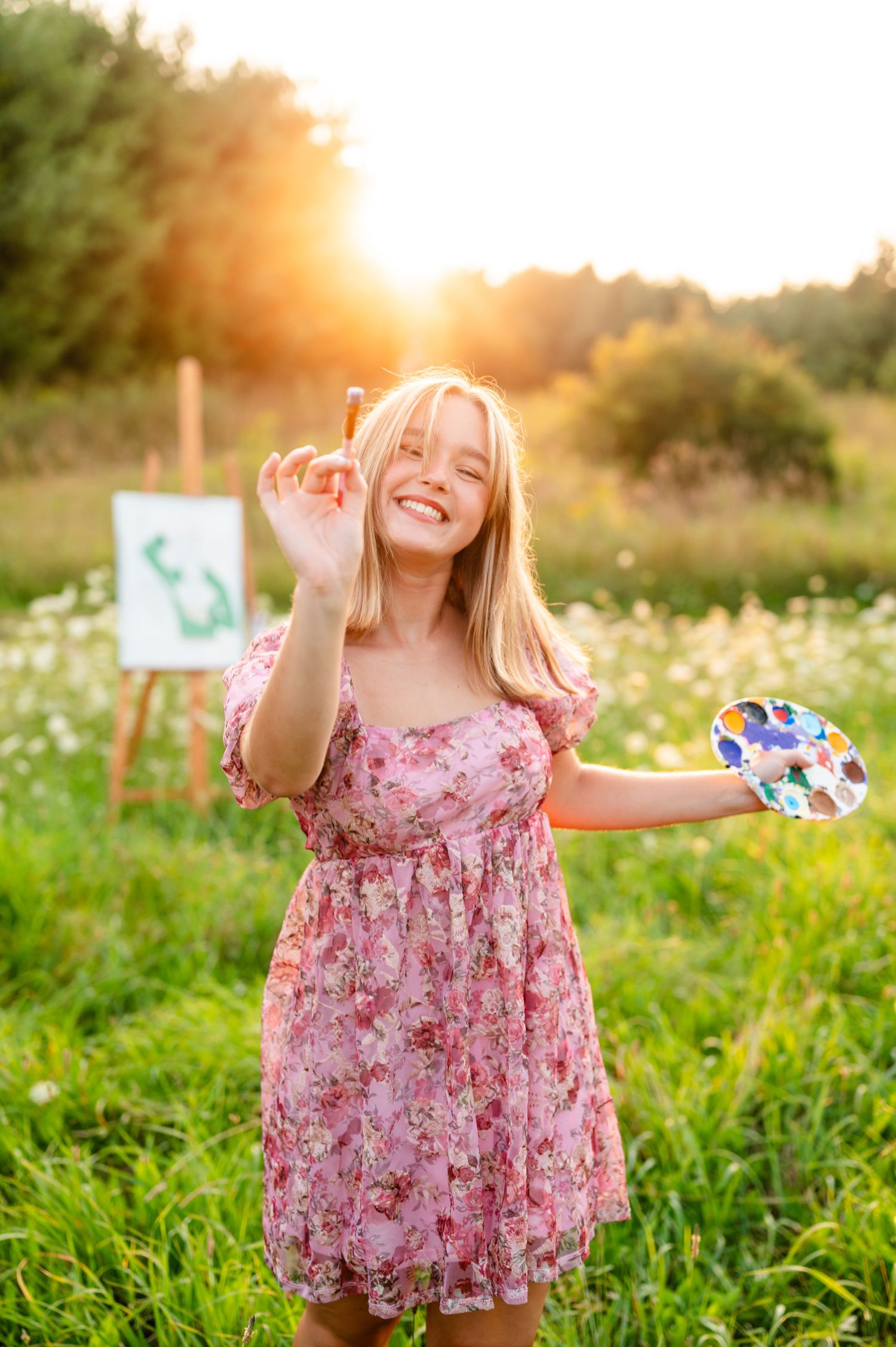Artist in floral pink dress holds paint palette and brush while standing in sunny wildflower meadow at sunset.