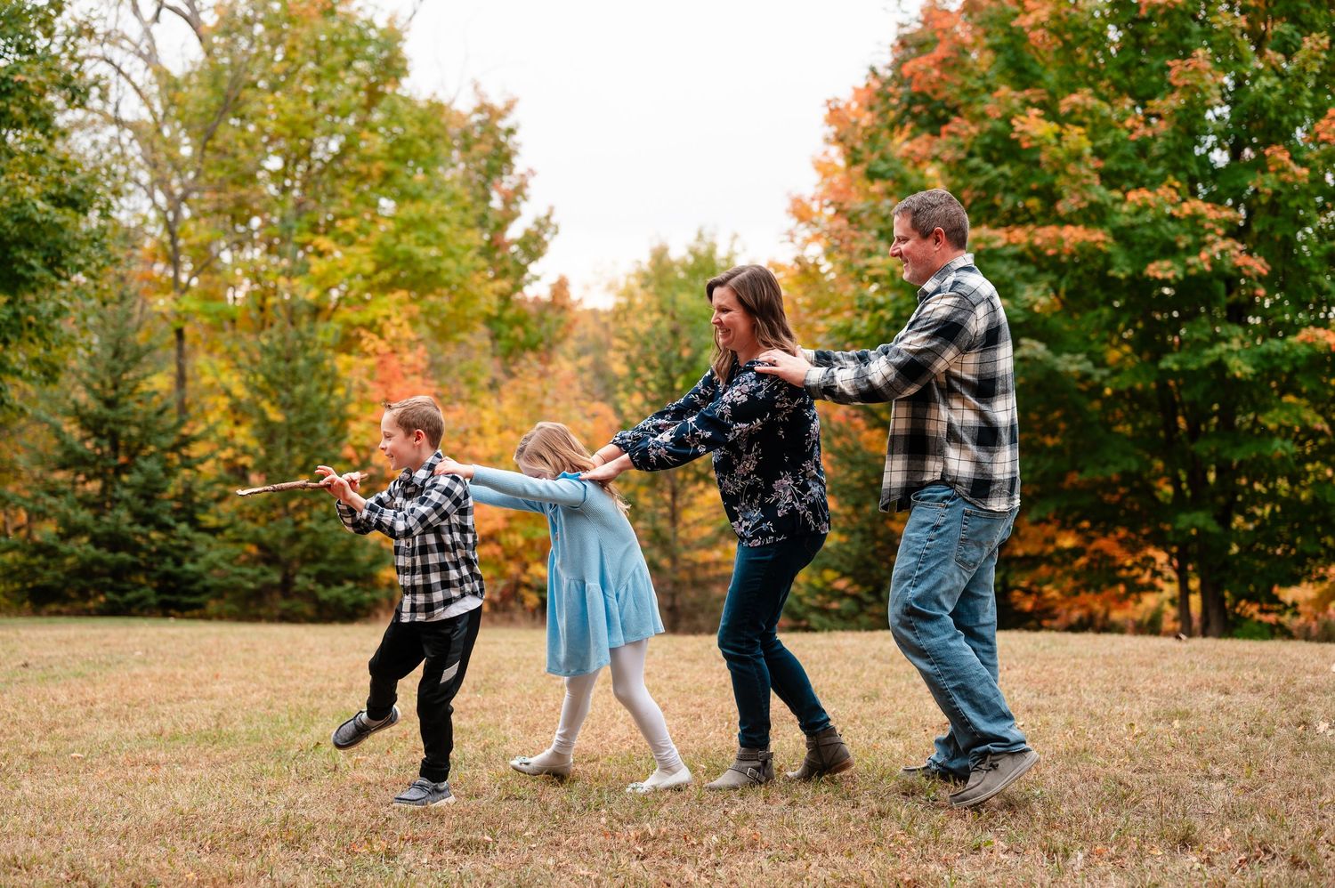 A family plays follow-the-leader in a field surrounded by colorful autumn trees in their backyard.