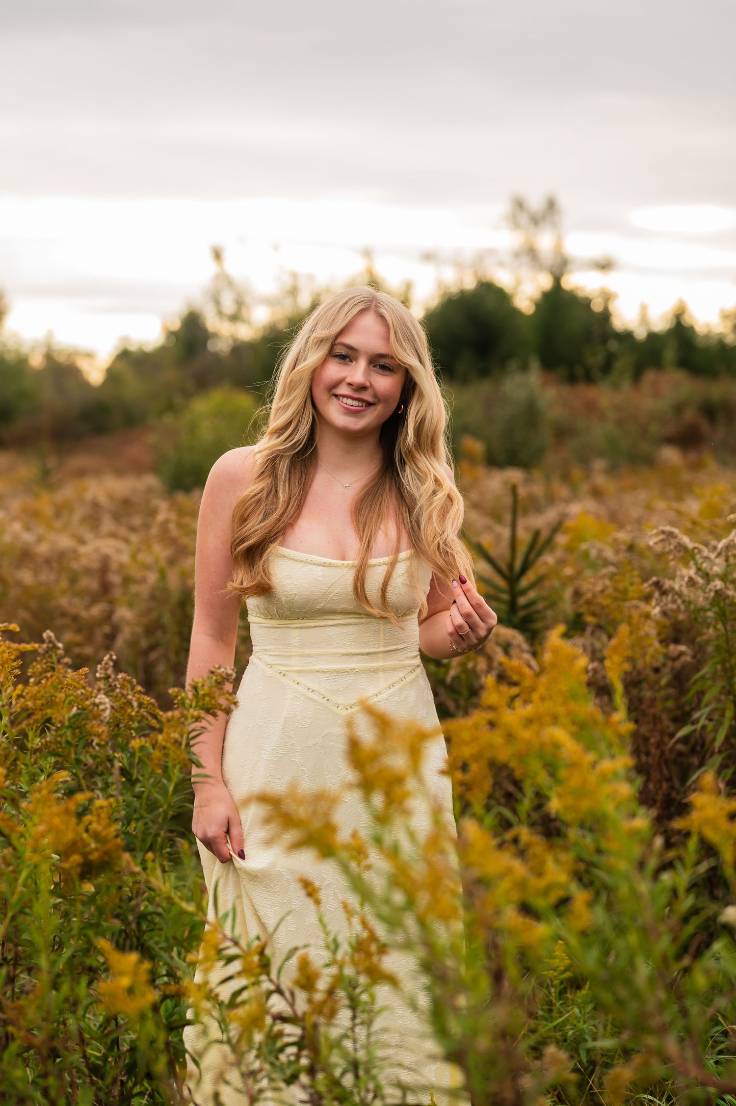 Senior girl in yellow dress standing among wildflowers at golden hour.