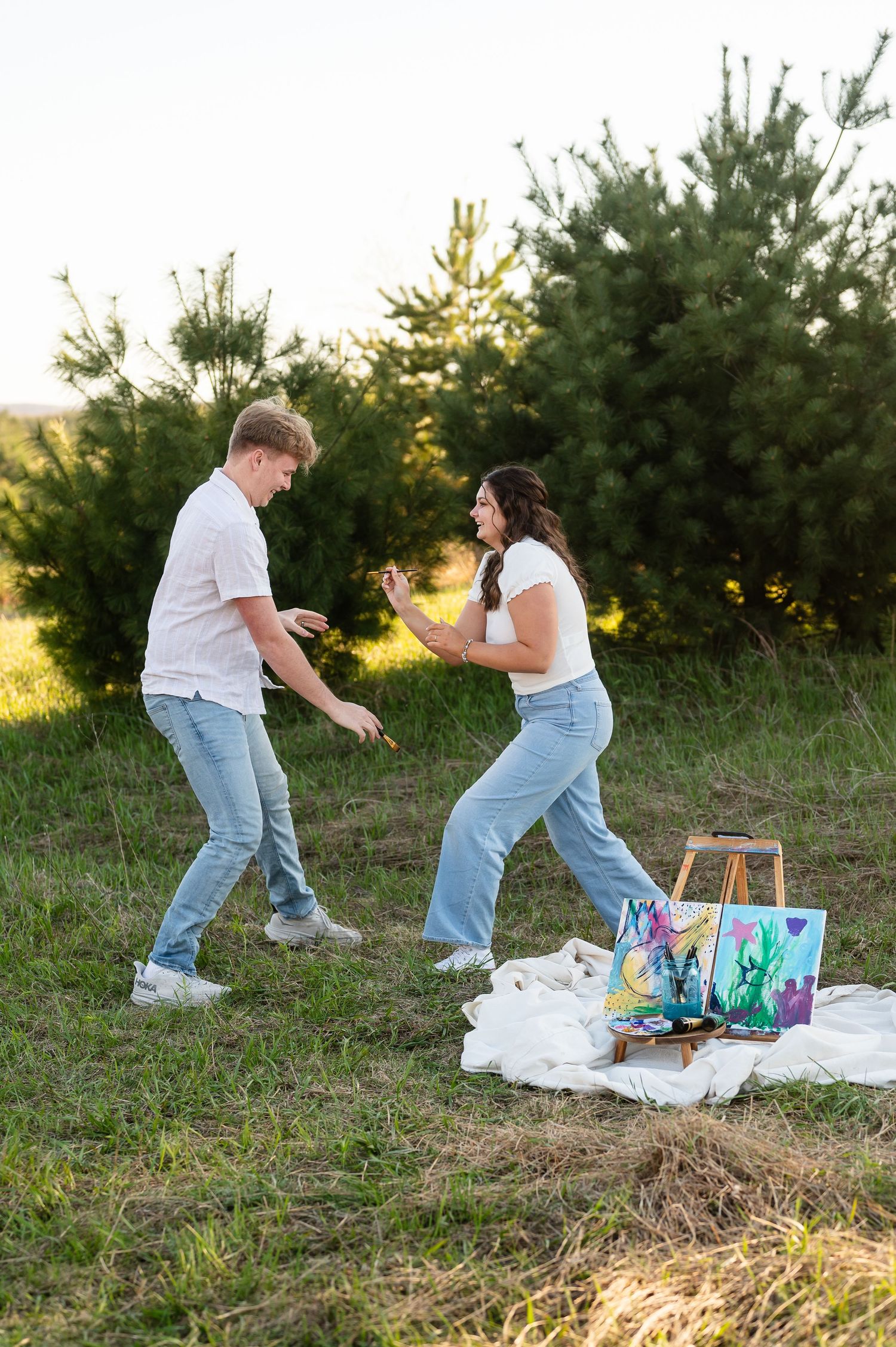 A playful couple dances together on a white blanket during an outdoor painting photoshoot.