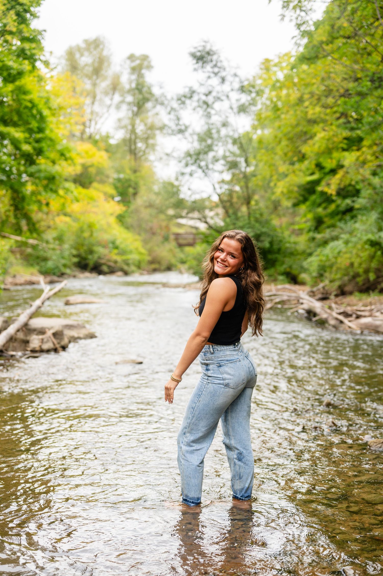 Senior girl walking through a shallow river wearing denim jeans on a sunny day looking back at the camera.