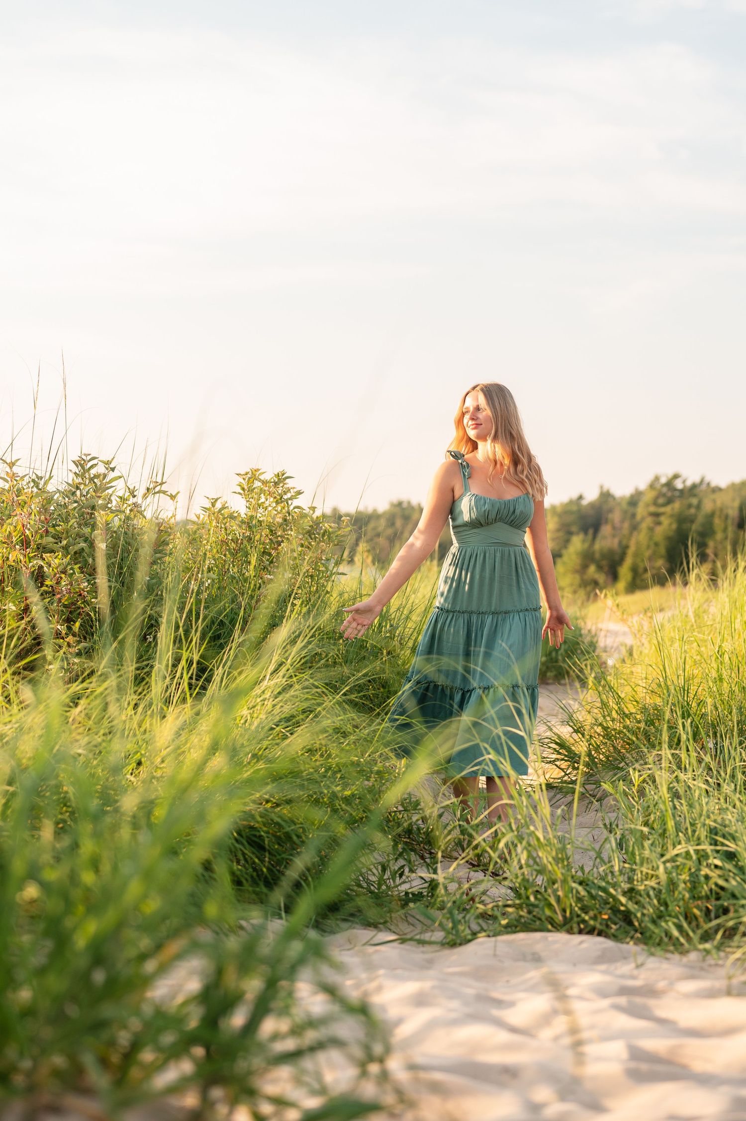 A senior girl in green dress walking among tall grass on a sandy beach at sunset in Northern Michigan.