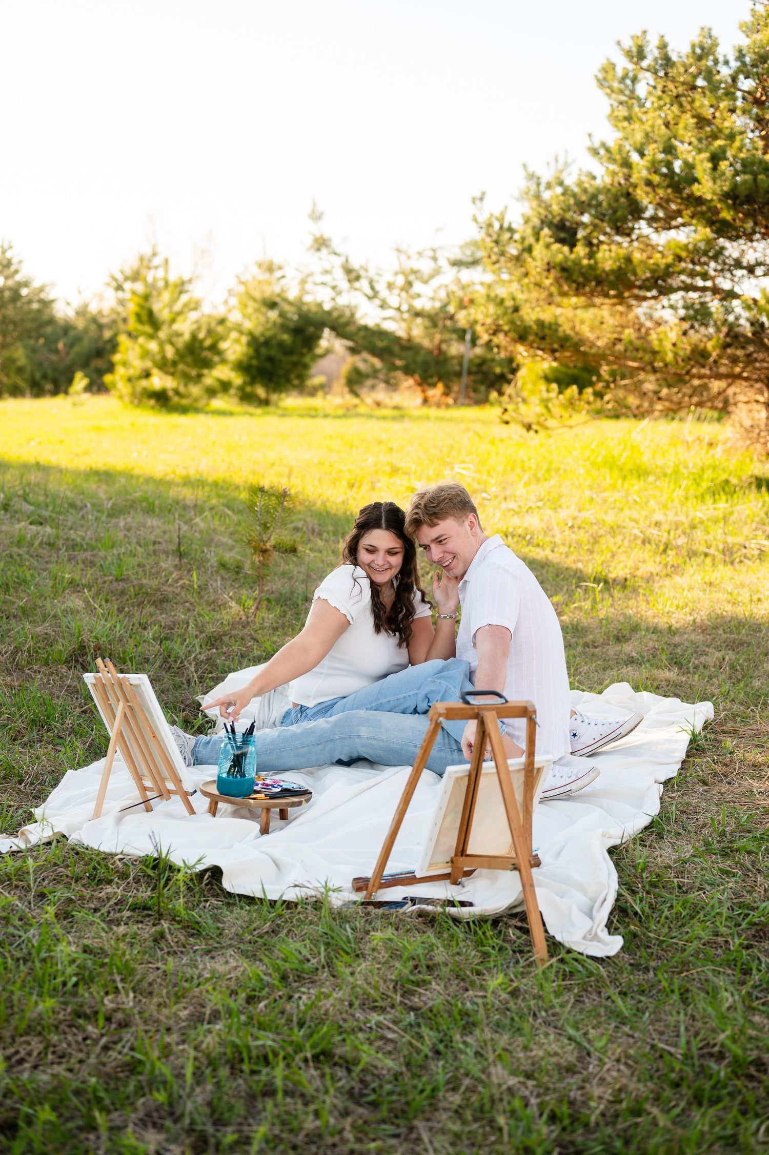 A couple relaxes on  a white blanket during an outdoor date admiring each others painting.