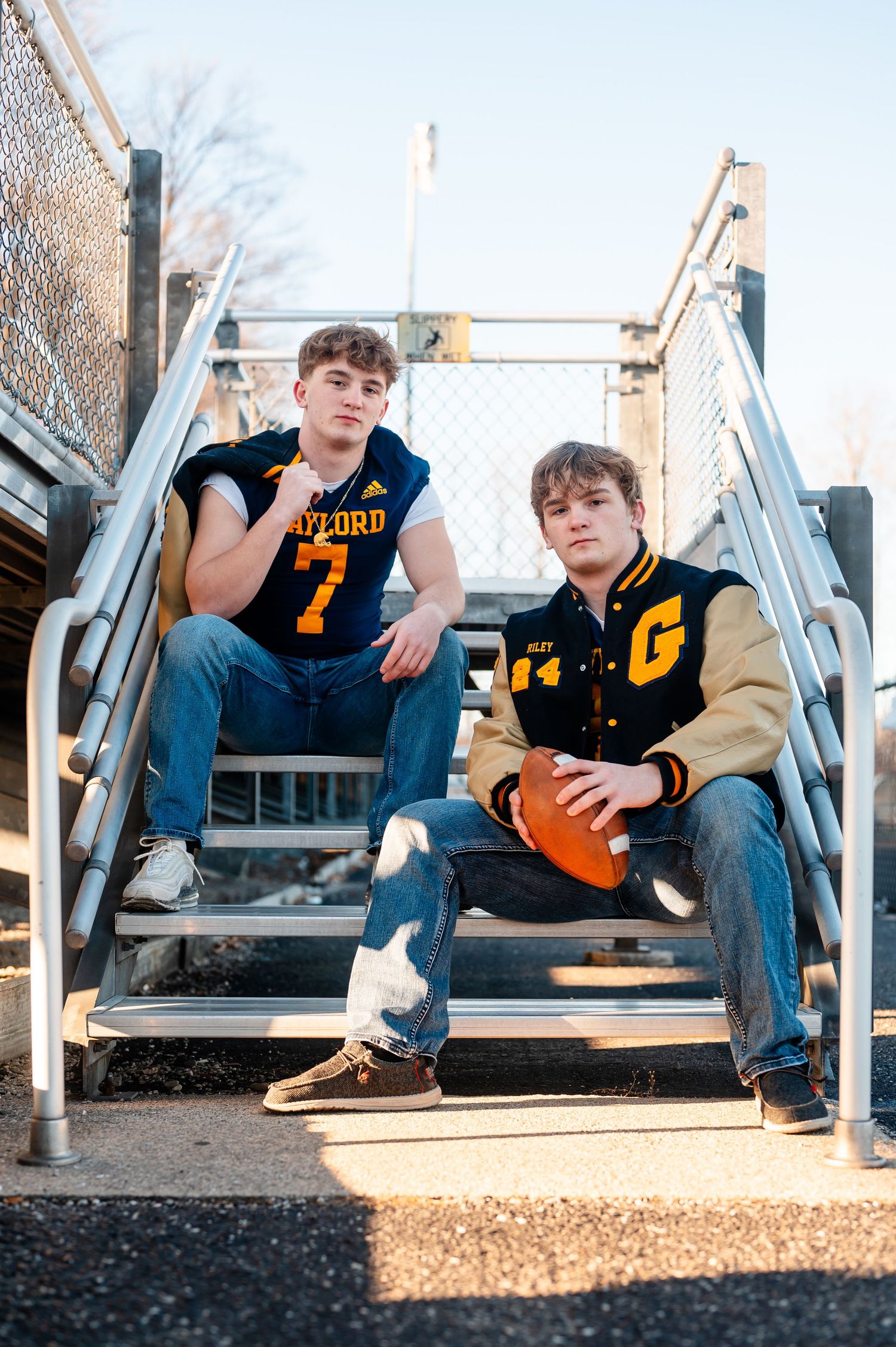 Senior boys in varsity jackets lounging on bleacher steps holding a football during golden hour.