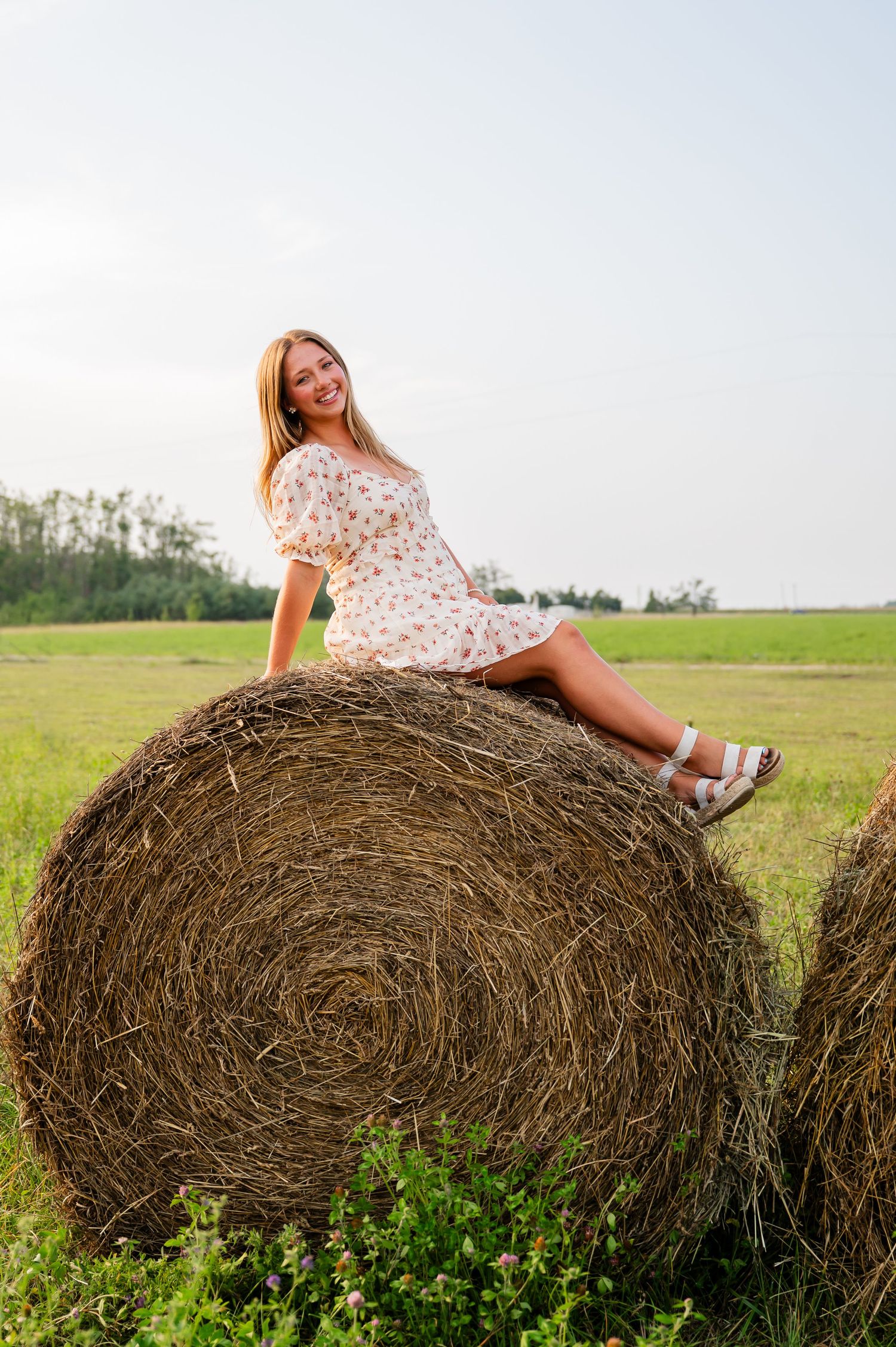 Person in floral dress poses playfully on hay bales in green field.