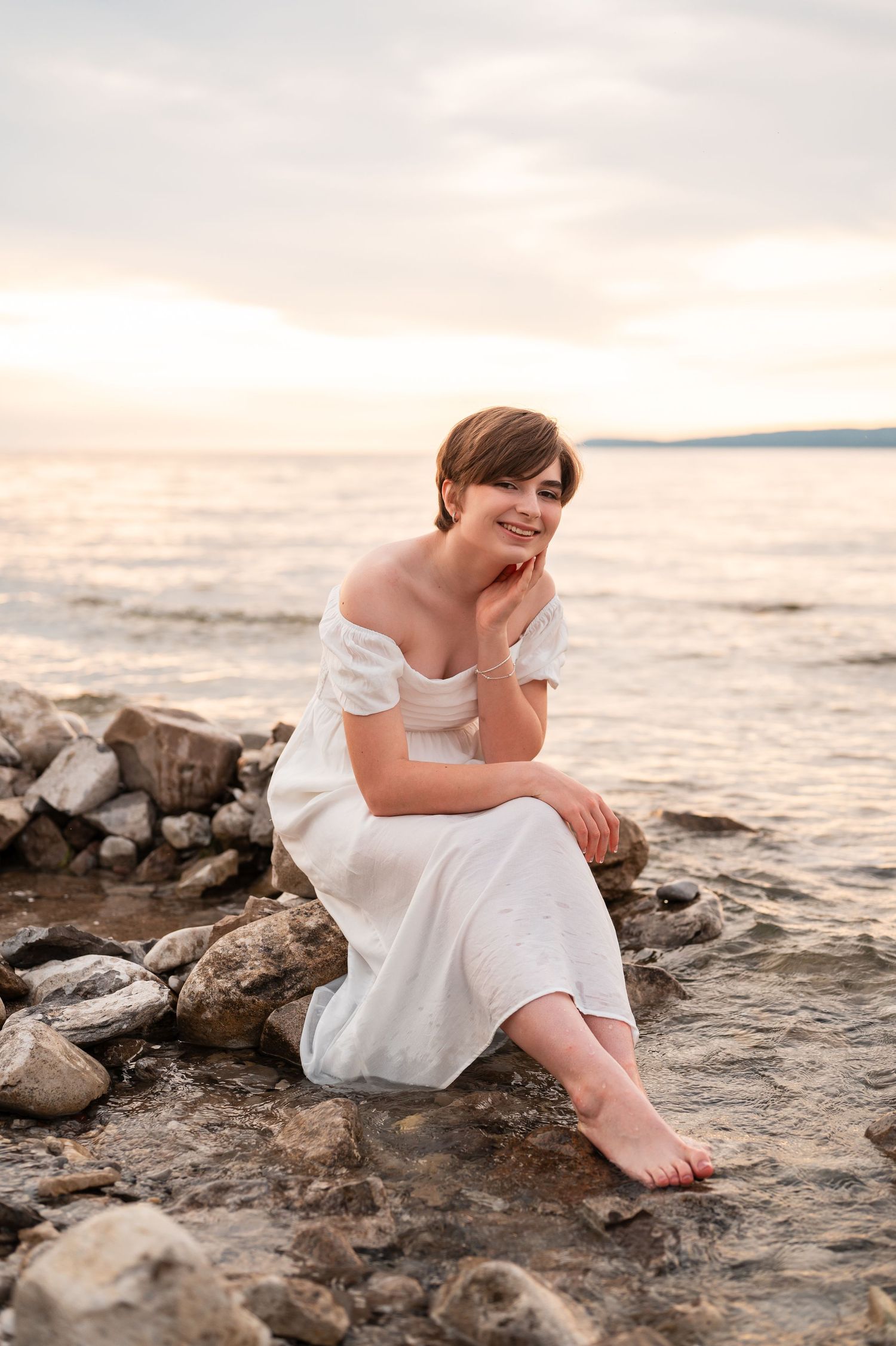 A senior girl in a white dress sits on rocky shoreline at sunset with dreamy skies.