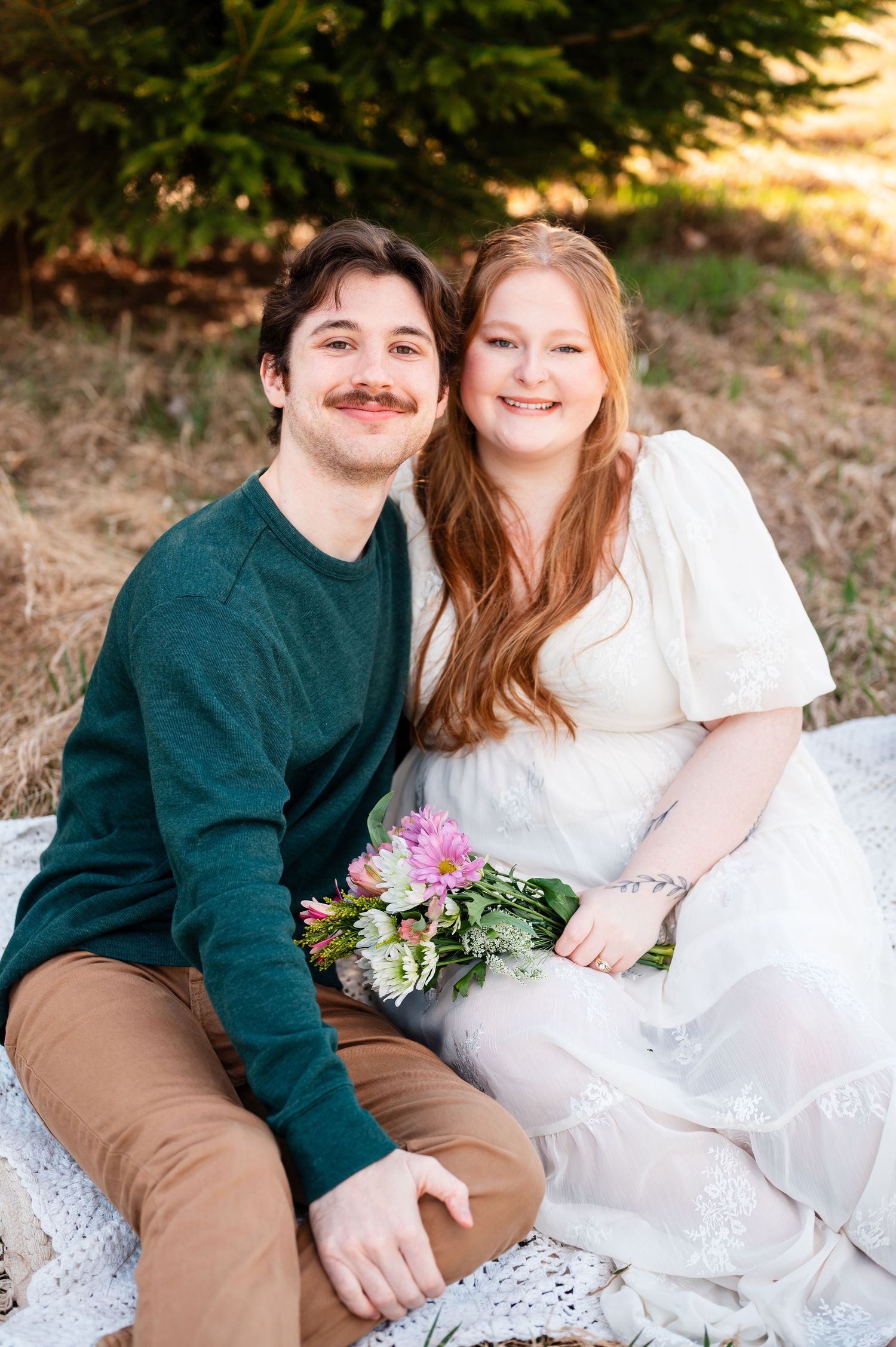 A couple in casual attire sits on a white blanket outdoors holding pink flowers.