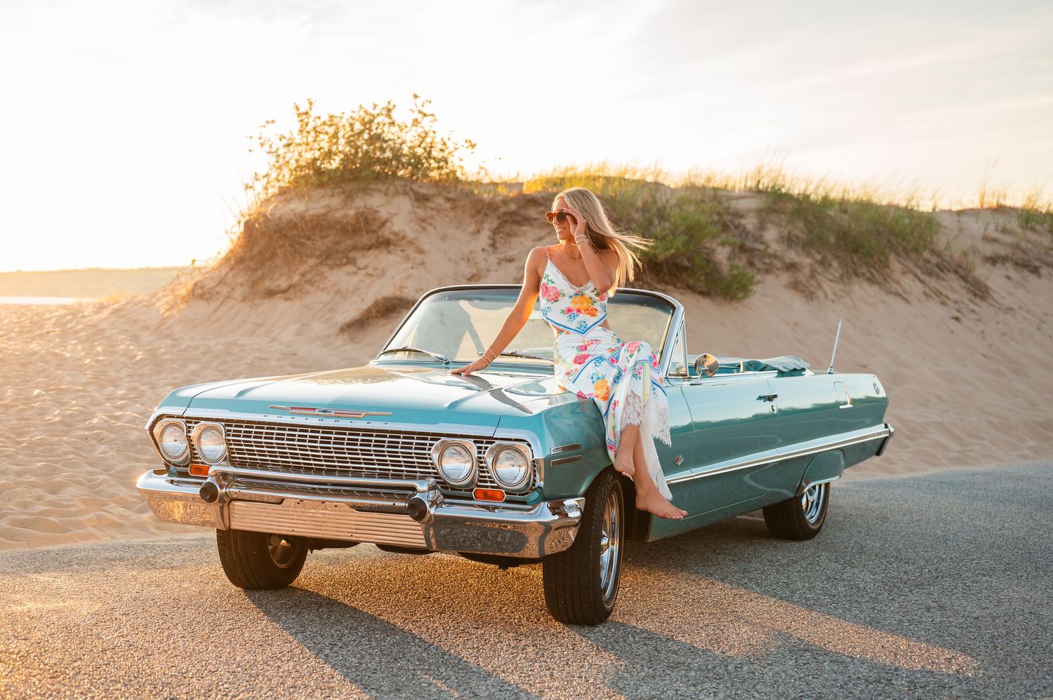 Woman in floral dress posing on a teal vintage convertible car near sandy dunes at golden sunset.