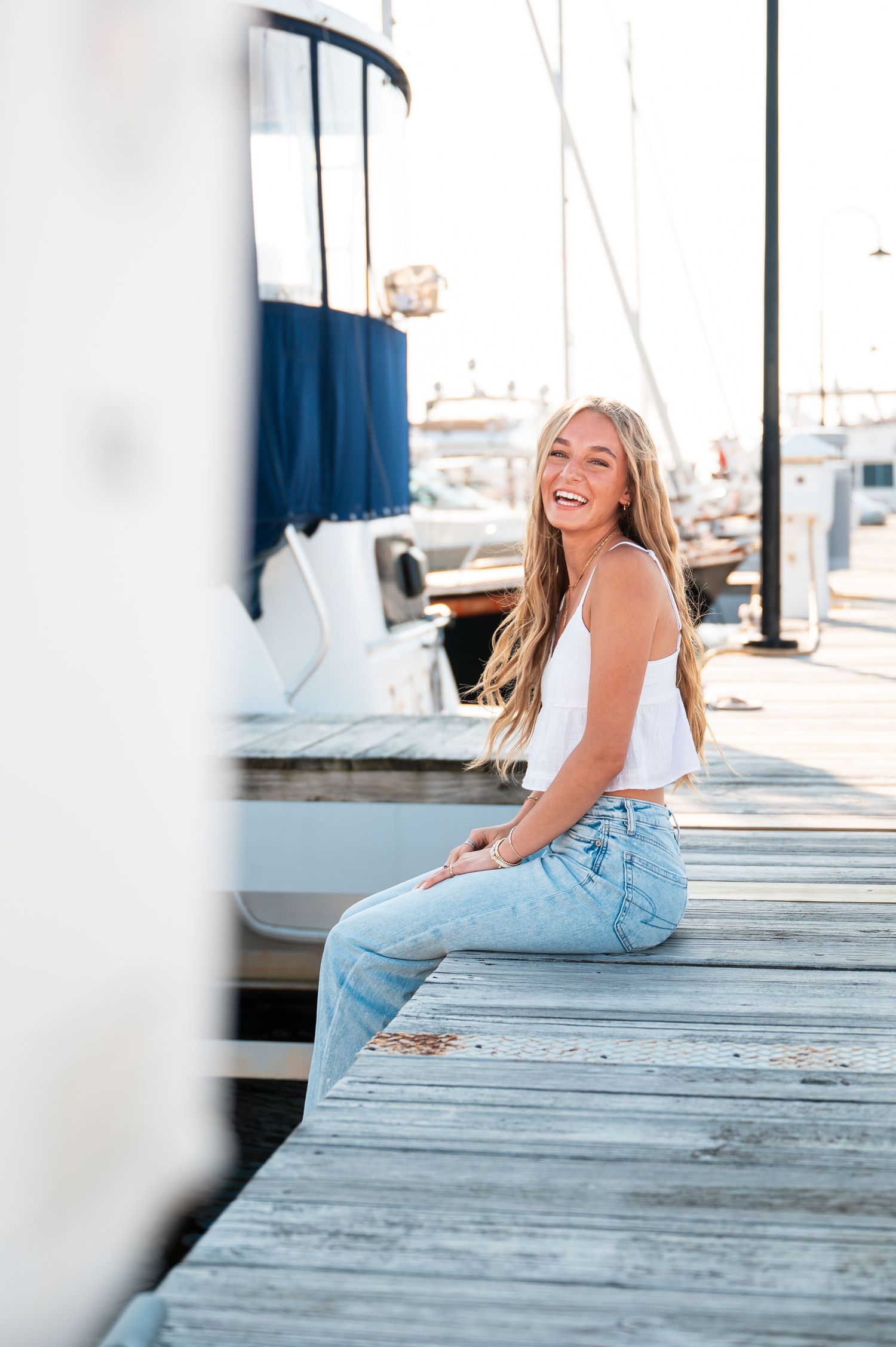 A blonde senior girl in casual attire sitting on wooden dock at marina on summers day.