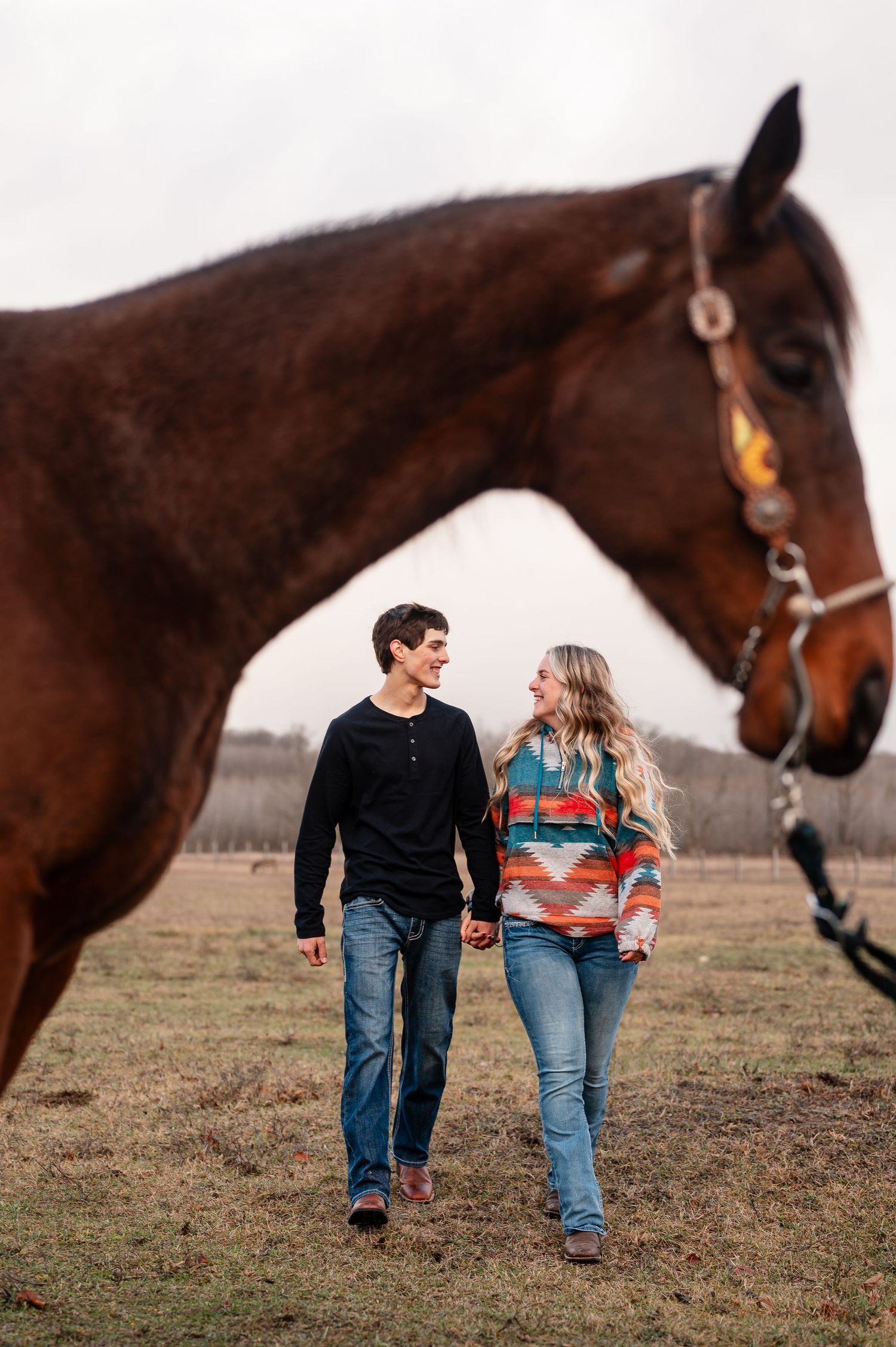 Couple walks together in field while brown horse follows behind them.