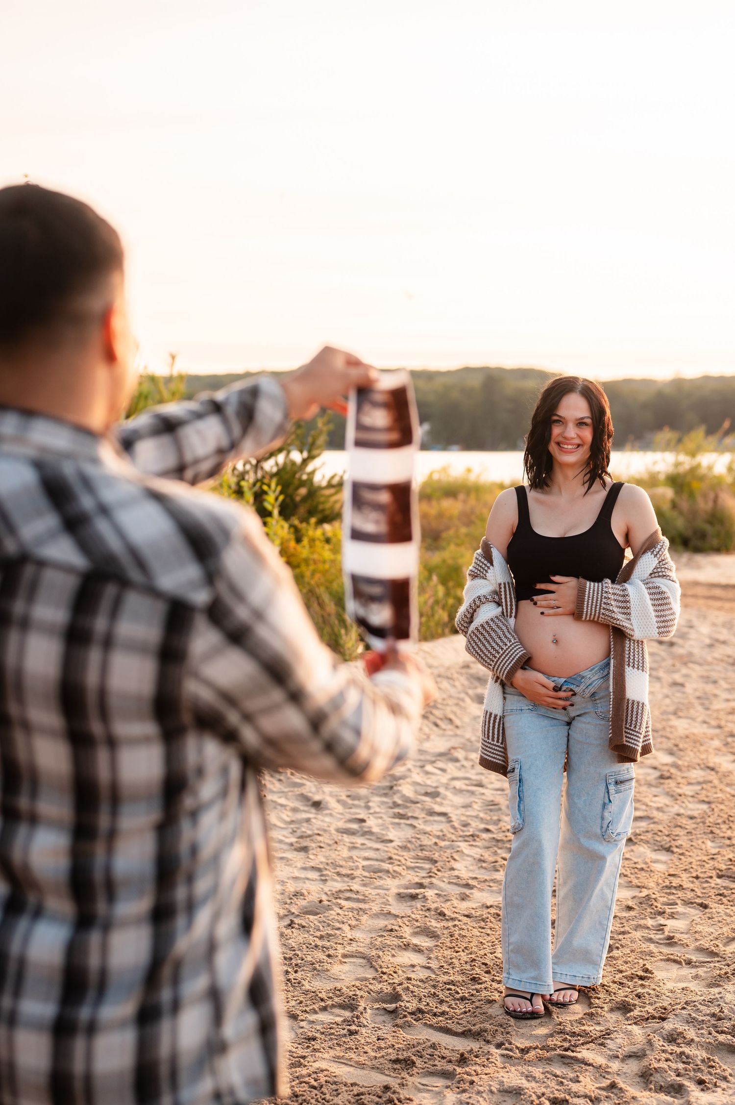 Couple sharing tender moments on a sandy beach at sunset wearing casual attire with ultrasound picture.