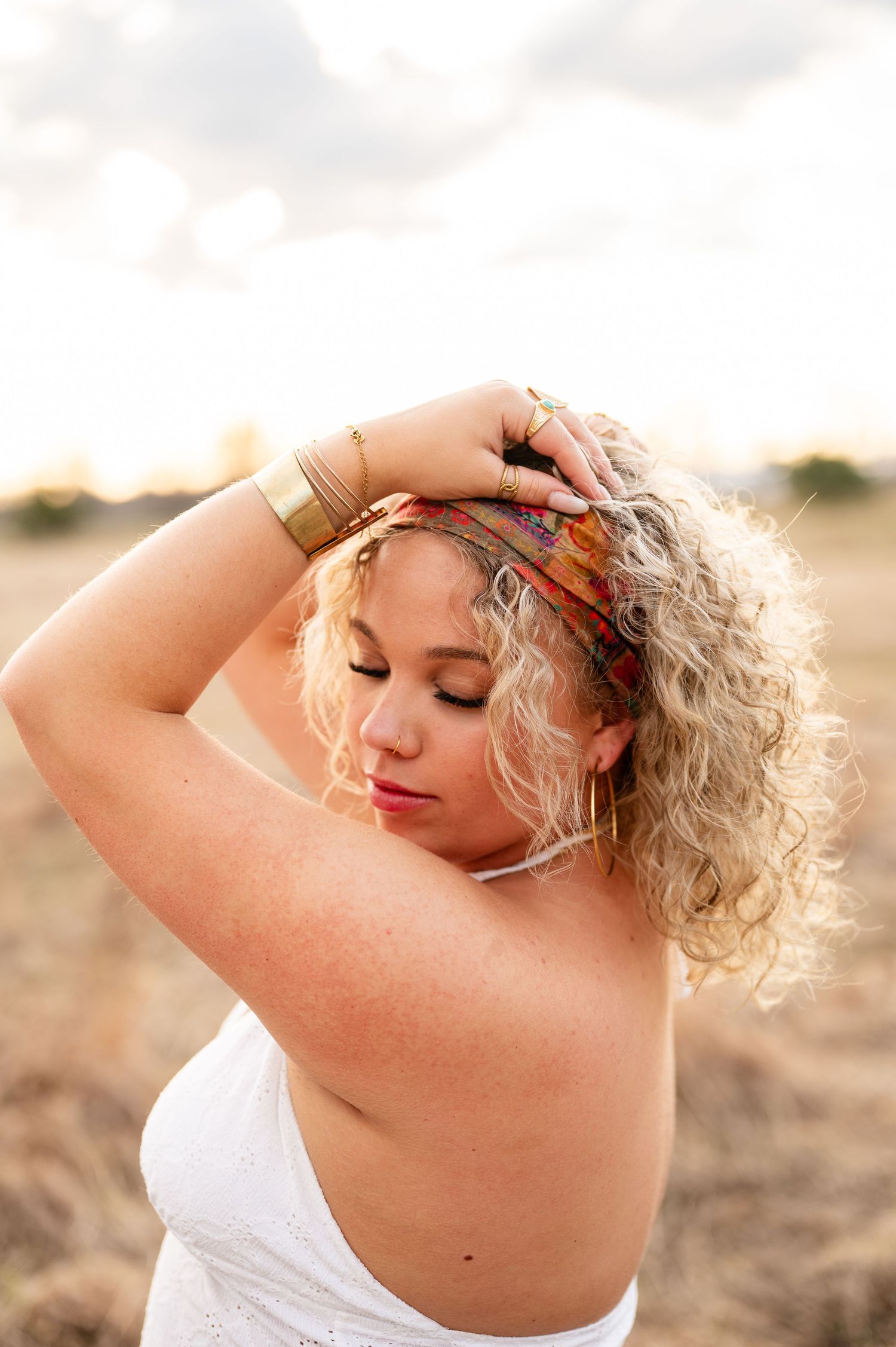 Curly blonde woman in white top wearing colorful headband and gold jewelry posing outdoors in golden field.