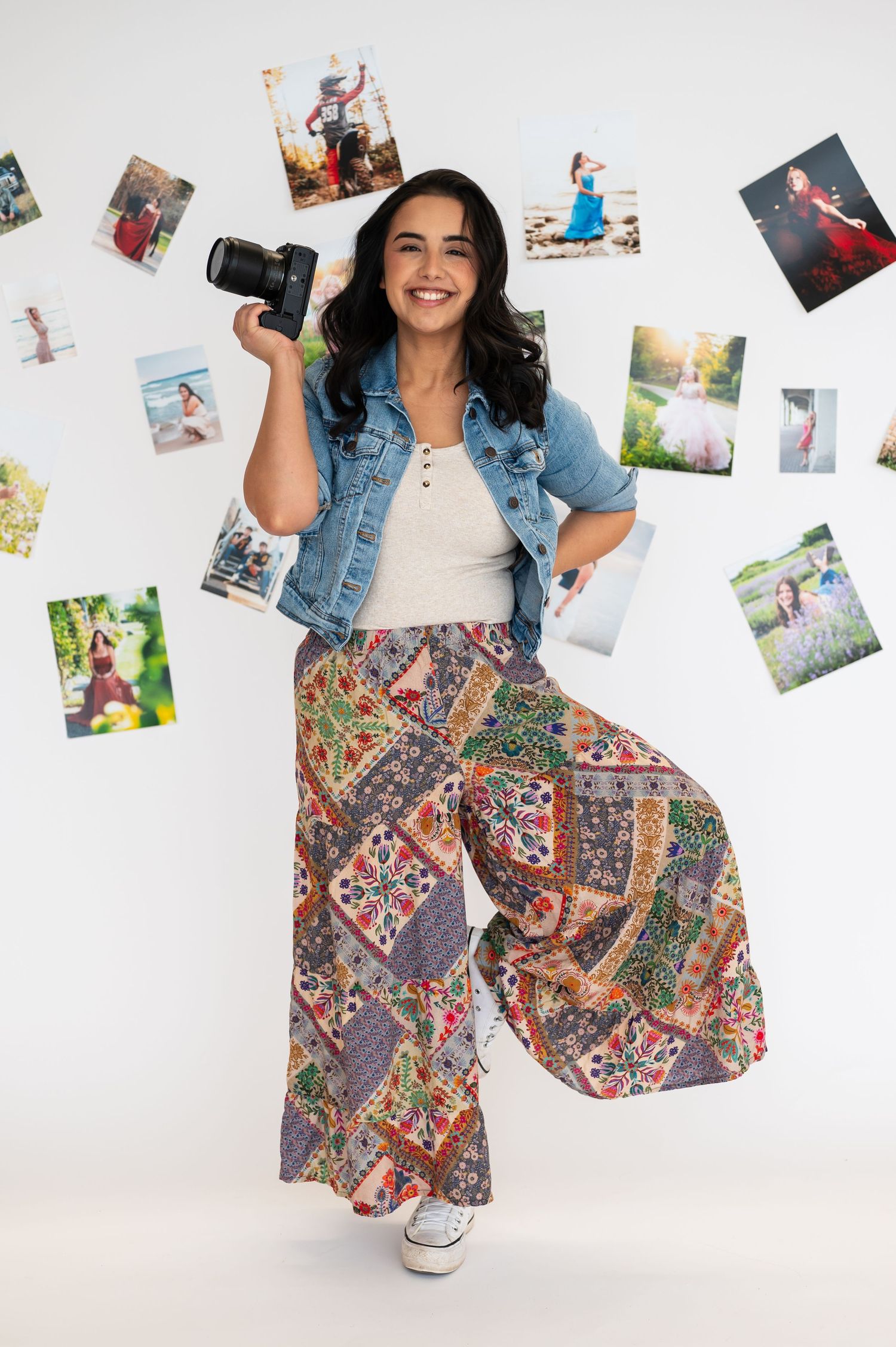 Photographer in denim jacket and patterned skirt poses with photos on white background.