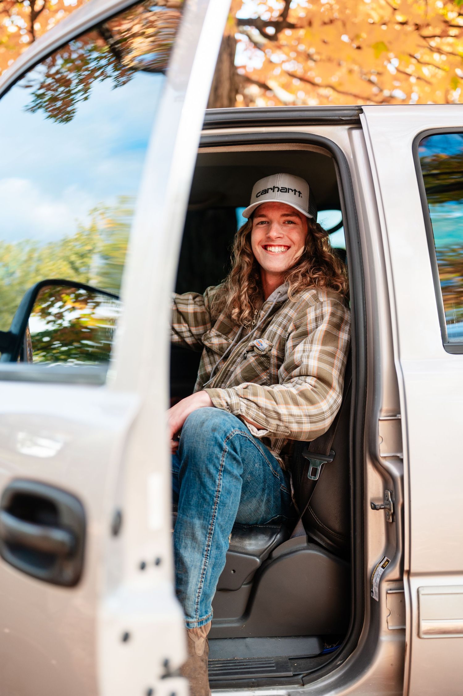 Senior boy in plaid shirt and baseball cap sitting in white truck during autumn season.