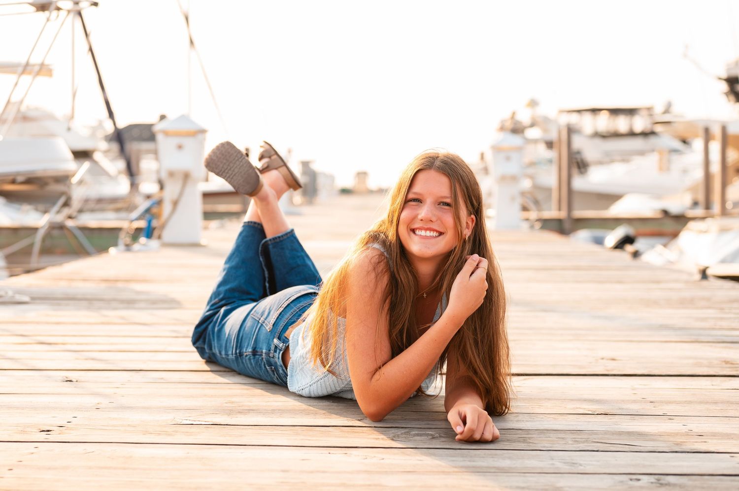 A senior girl relaxing on a wooden dock by the water during sunset in a light blue top and jeans.
