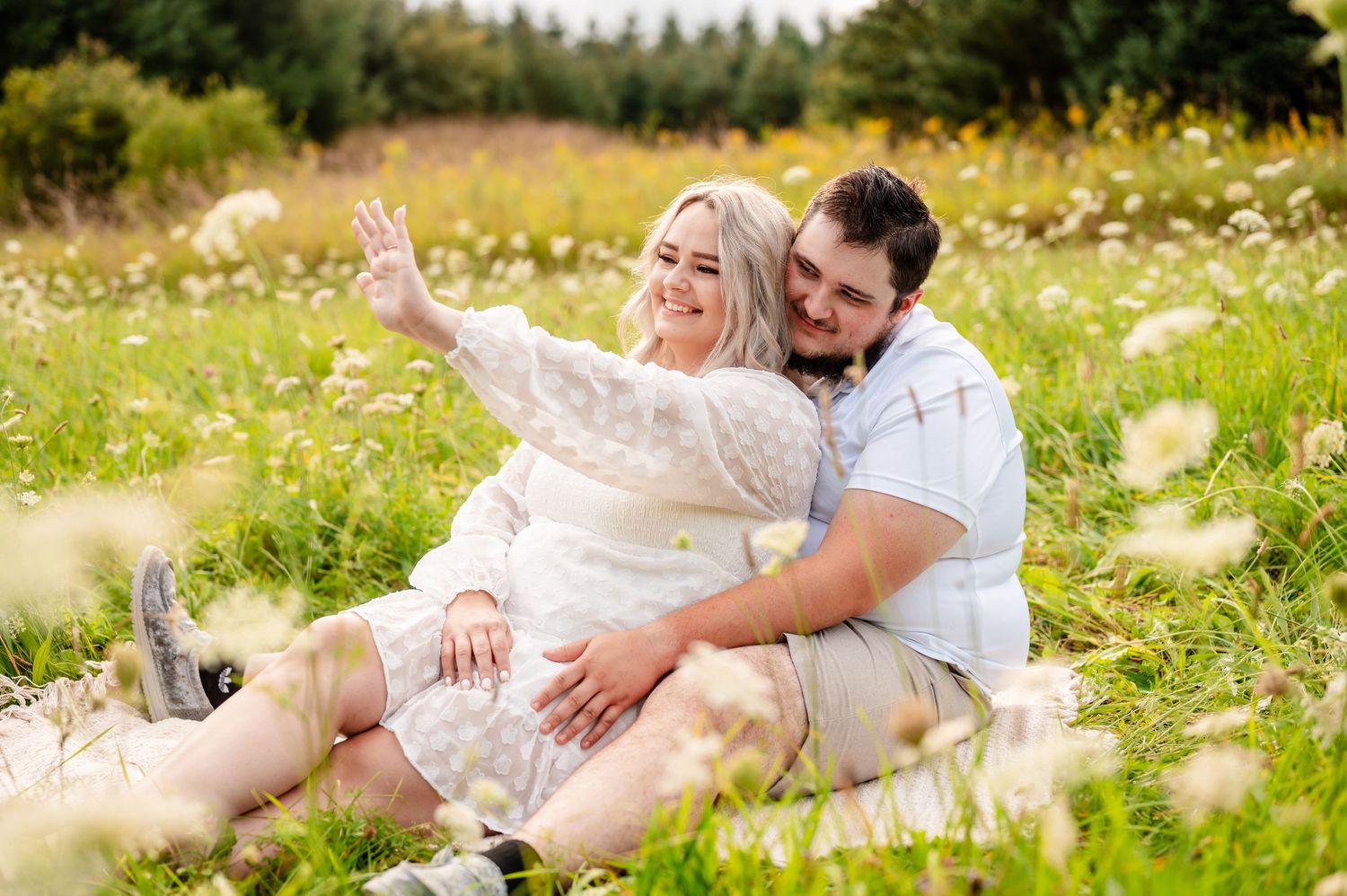 Couple sitting around in a sunny wildflower meadow with tall grass and trees in the background admiring an engagement ring together.