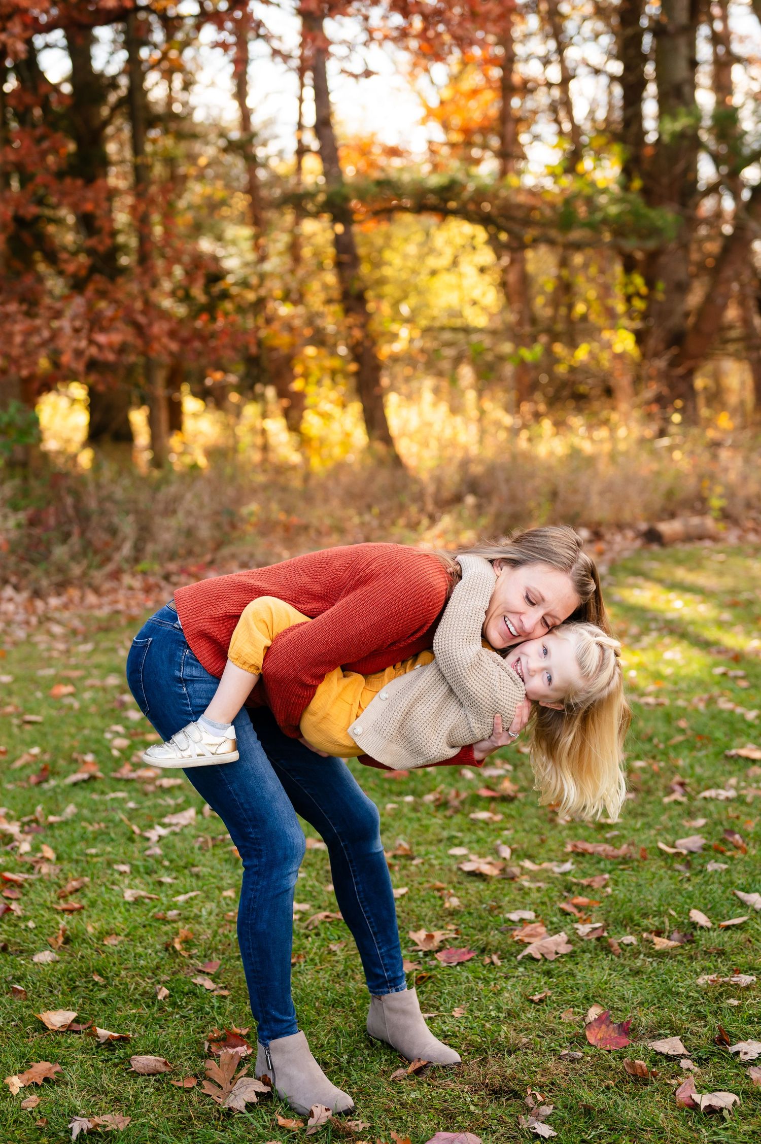 Playful scene of mother and daughter bending over in autumn leaves during outdoor photo session.