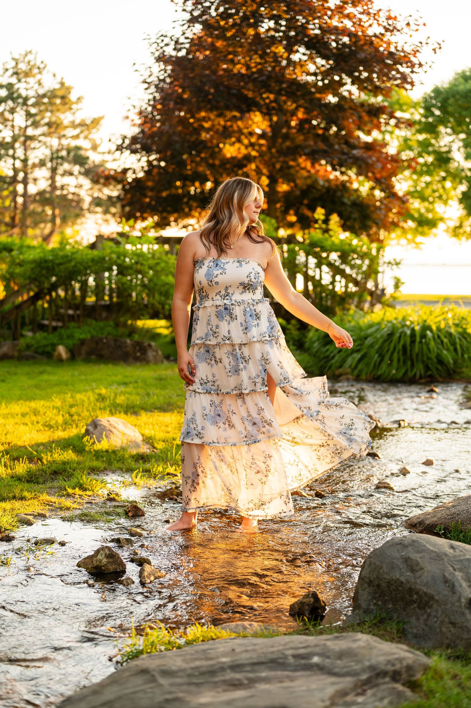 Woman in floral tiered maxi dress wading through a rocky stream at golden hour sunset in a lush garden.