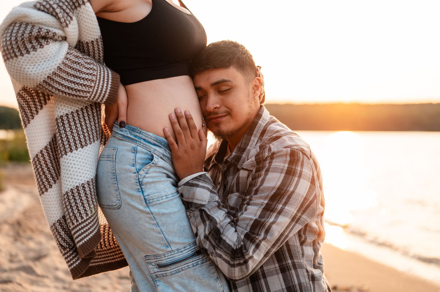 Maternity beach photoshoot at sunset with couple in casual clothing embracing.