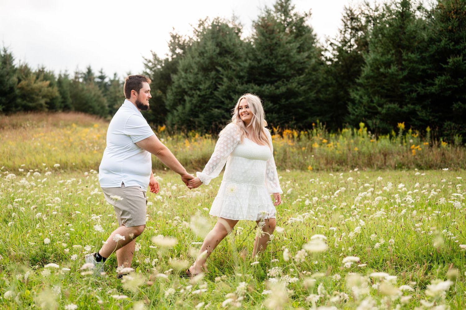 Couple laughing and playing together in a sunlit field of white wildflowers and tall grass.