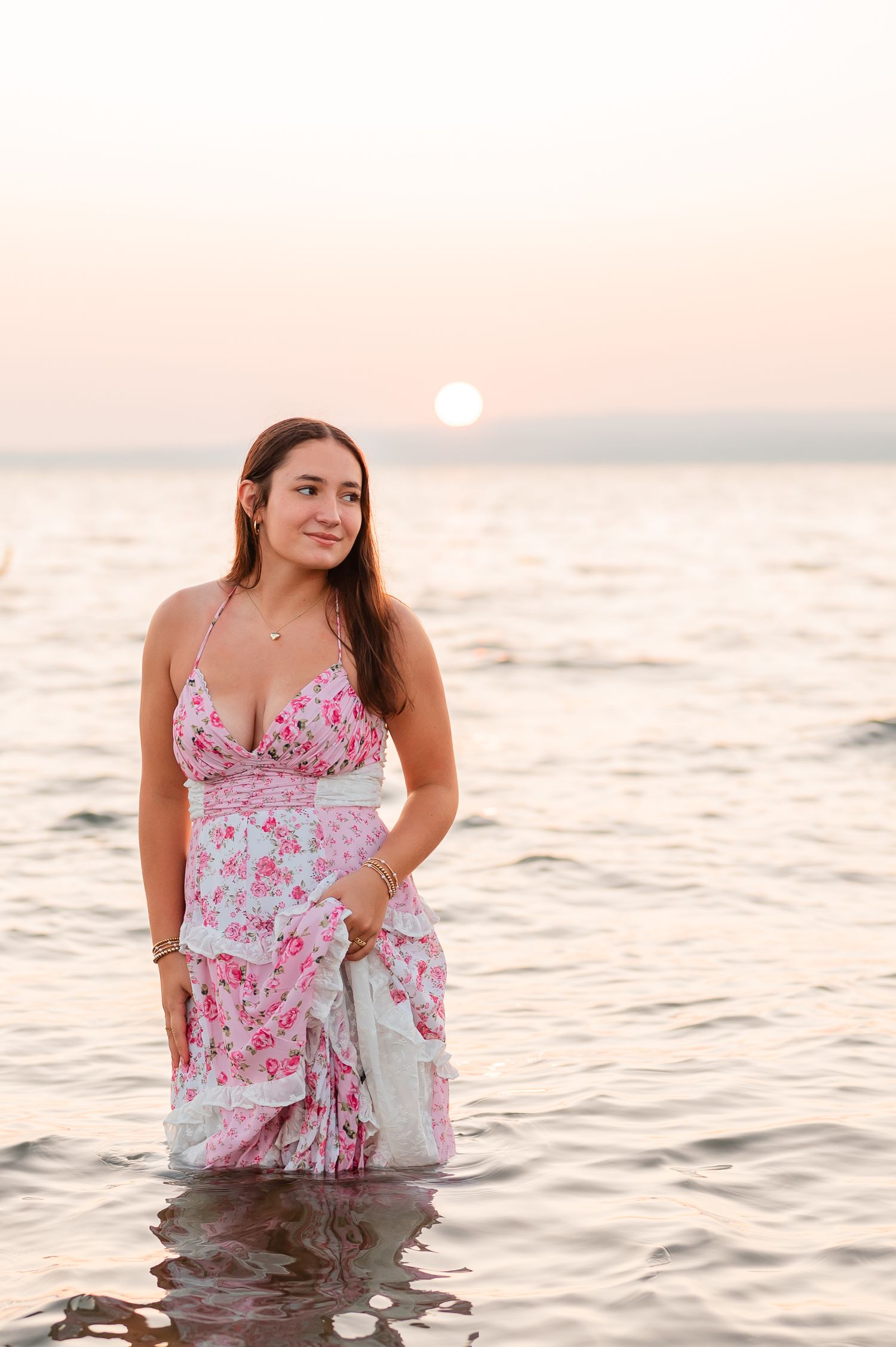A senior girl in a pink floral dress poses in shallow ocean water during sunset creating a dreamy beach portrait.