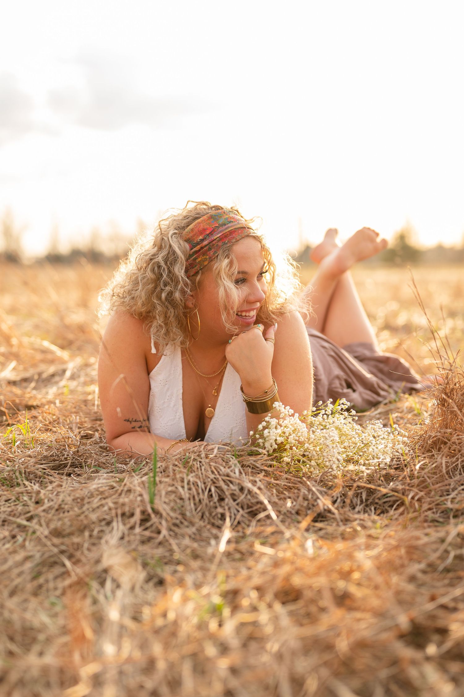 Smiling curly-haired woman lying in golden field at sunset, wearing white top and headband, holding wildflowers.