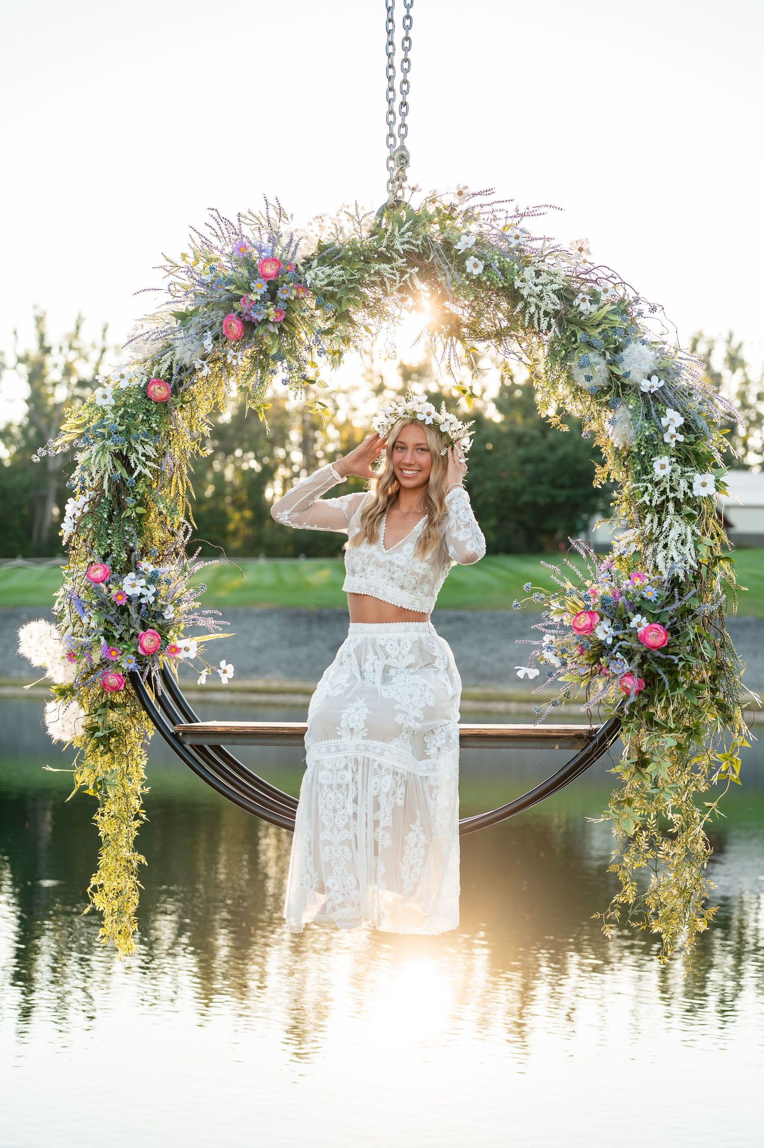 Woman in white lace outfit stands on floral hoop swing over water at golden hour with flower crown.