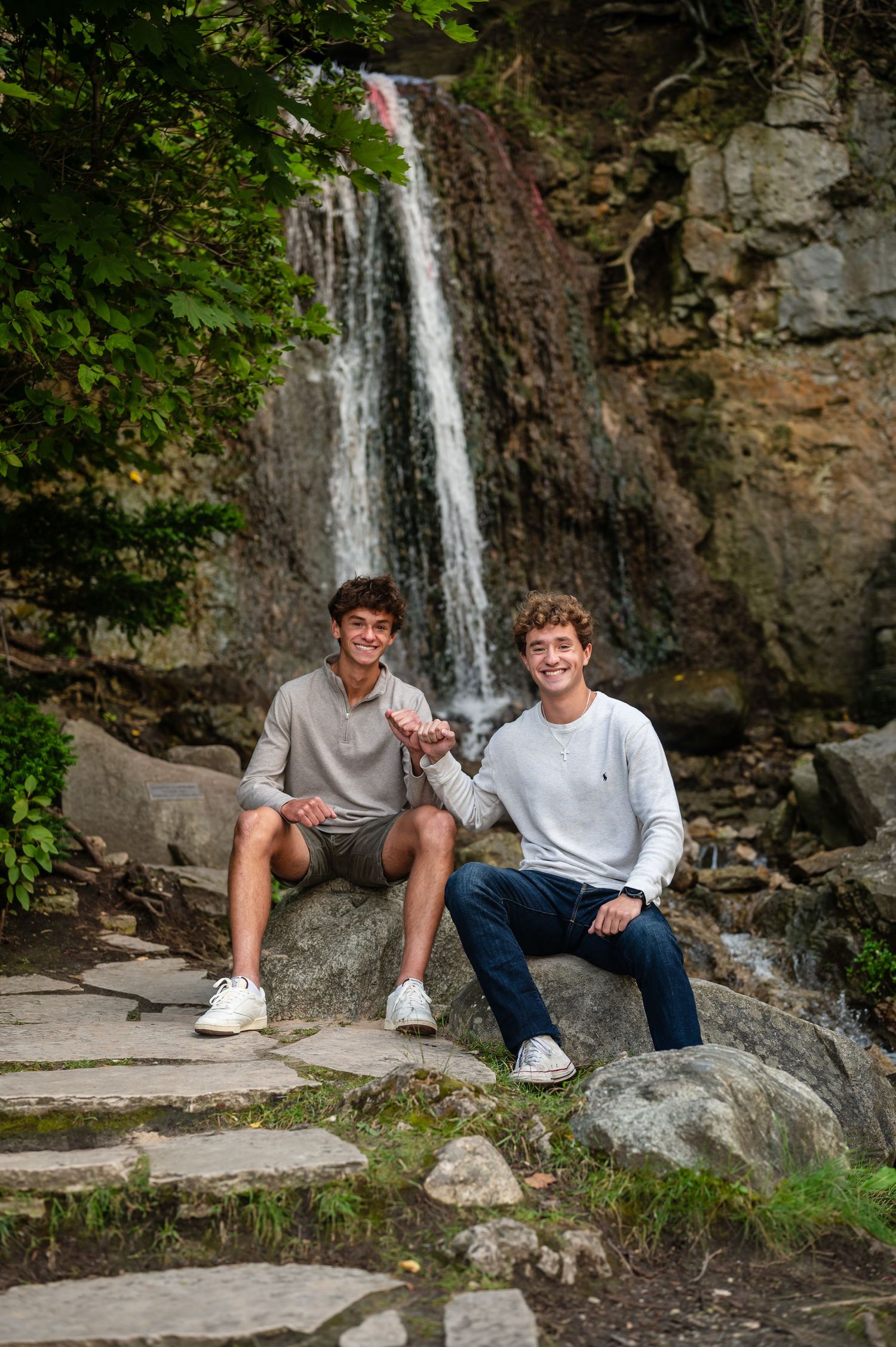 Two twin brothers sitting on rocks near scenic waterfall flowing as they smile.